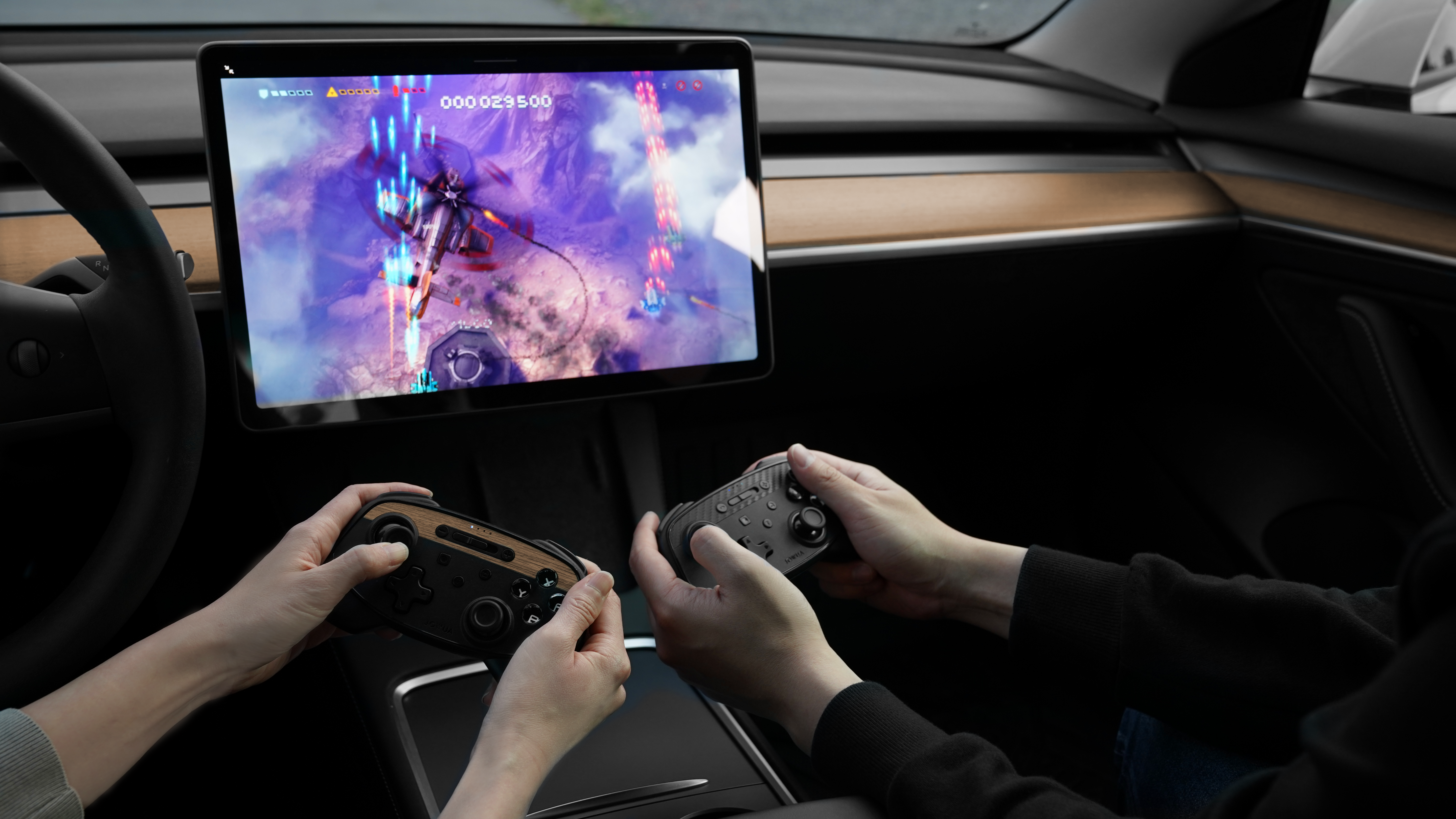 兩個人坐在車內,手持視訊遊戲控制器,玩著顯示在車內中央觸控螢幕上的彩色動作遊戲。車內裝潢充滿現代感,配備流線型儀表板和木質飾邊。.