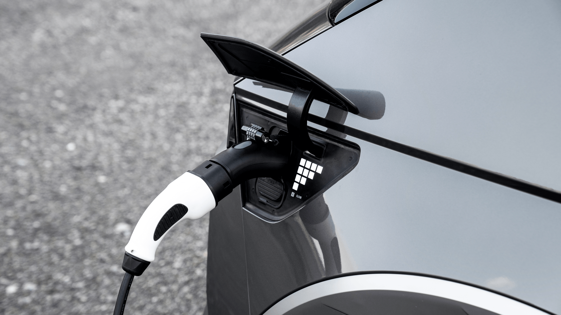特寫一輛正在充電的電動車,充電線已插入車輛的充電埠。這輛車是銀色的,停在灰色的砂石路面上。.