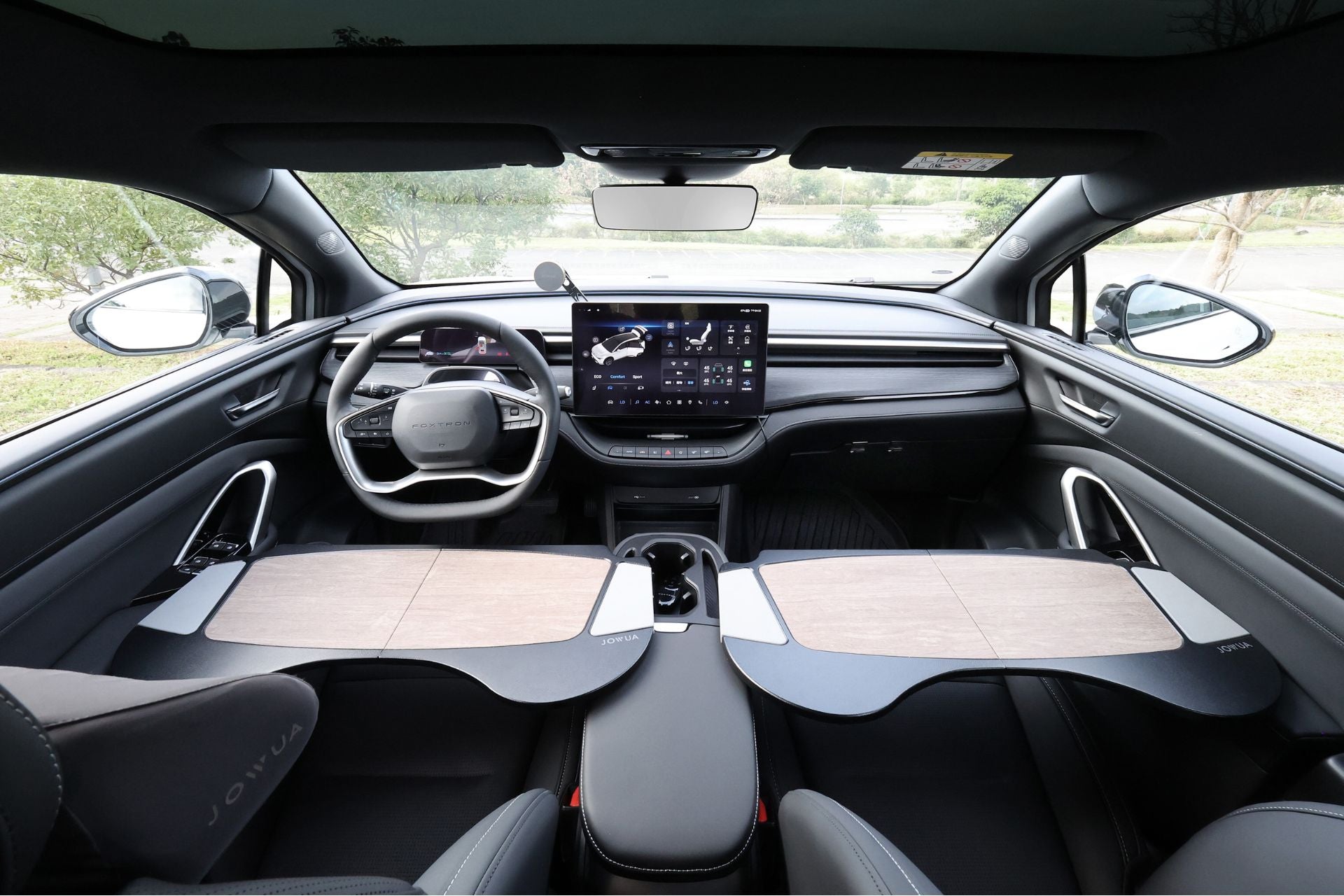 現代電動車的內視圖,顯示極簡的儀表板,配備大型中央觸控螢幕、未來感十足的方向盤,以及前座上方的兩張摺疊式木桌。.