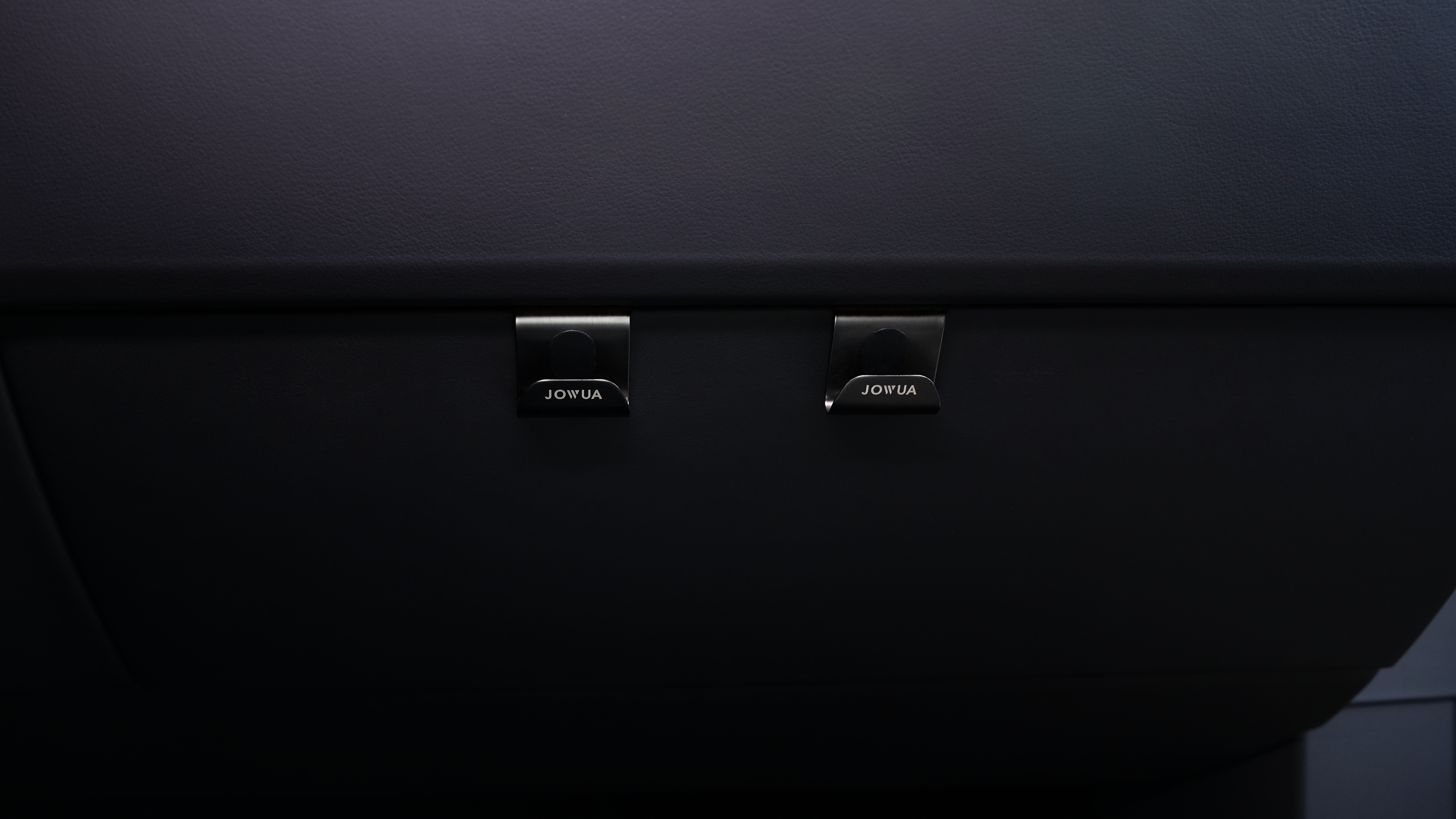 深色表面的特寫,顯示兩個帶有“JOWUA Taiwan”標籤的小掛鉤,專為 Model S/X 和 Model 3/Y 車輛設計。時尚的設計與光滑、中性的面板背景完美融合,相得益彰。.