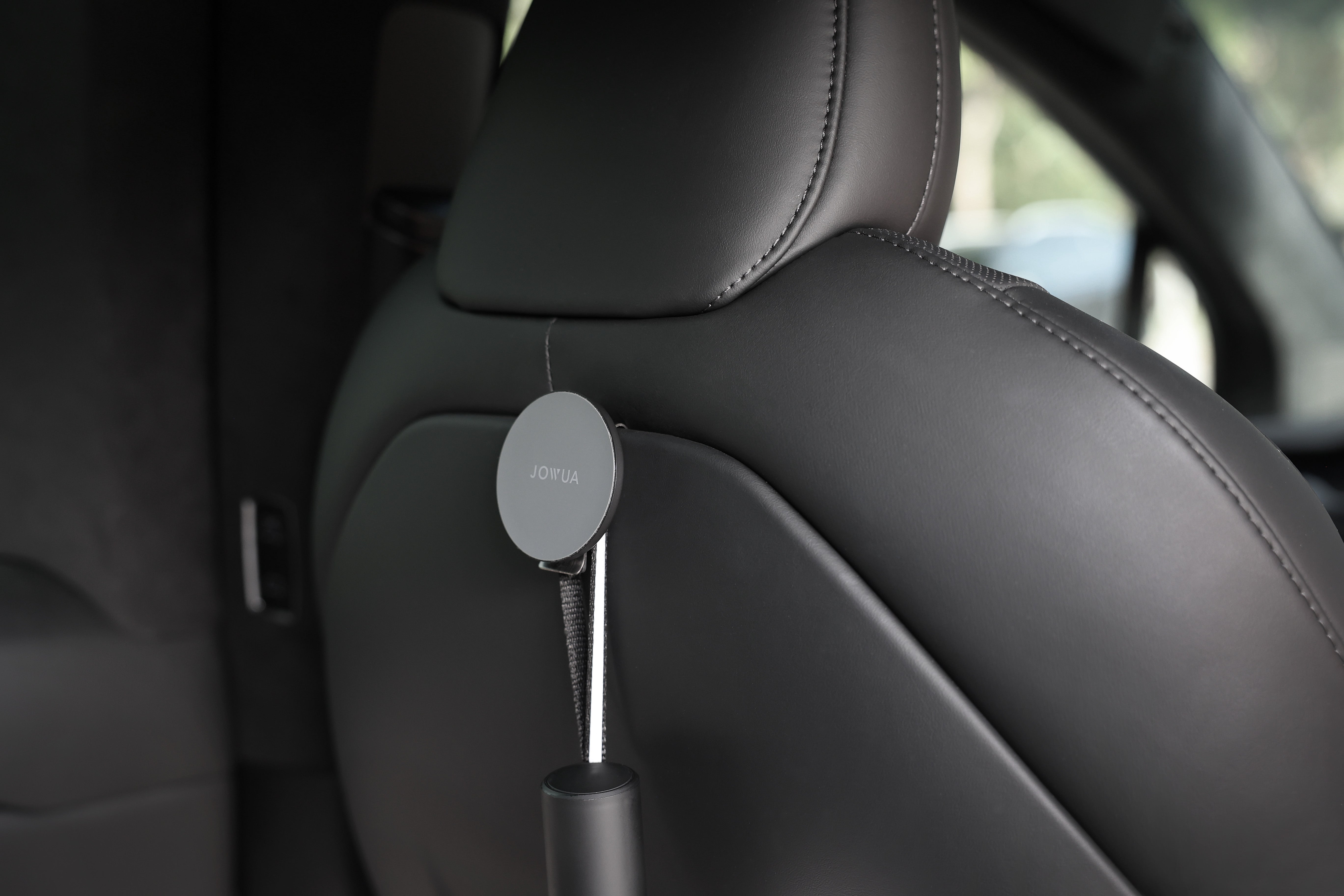 JOWUA台灣MagSafe磁吸掛鉤組(Model S/X/Y煥新版)附在車內黑色Model Y椅背頭枕上的特寫,透過車窗看到模糊的綠色植物。.