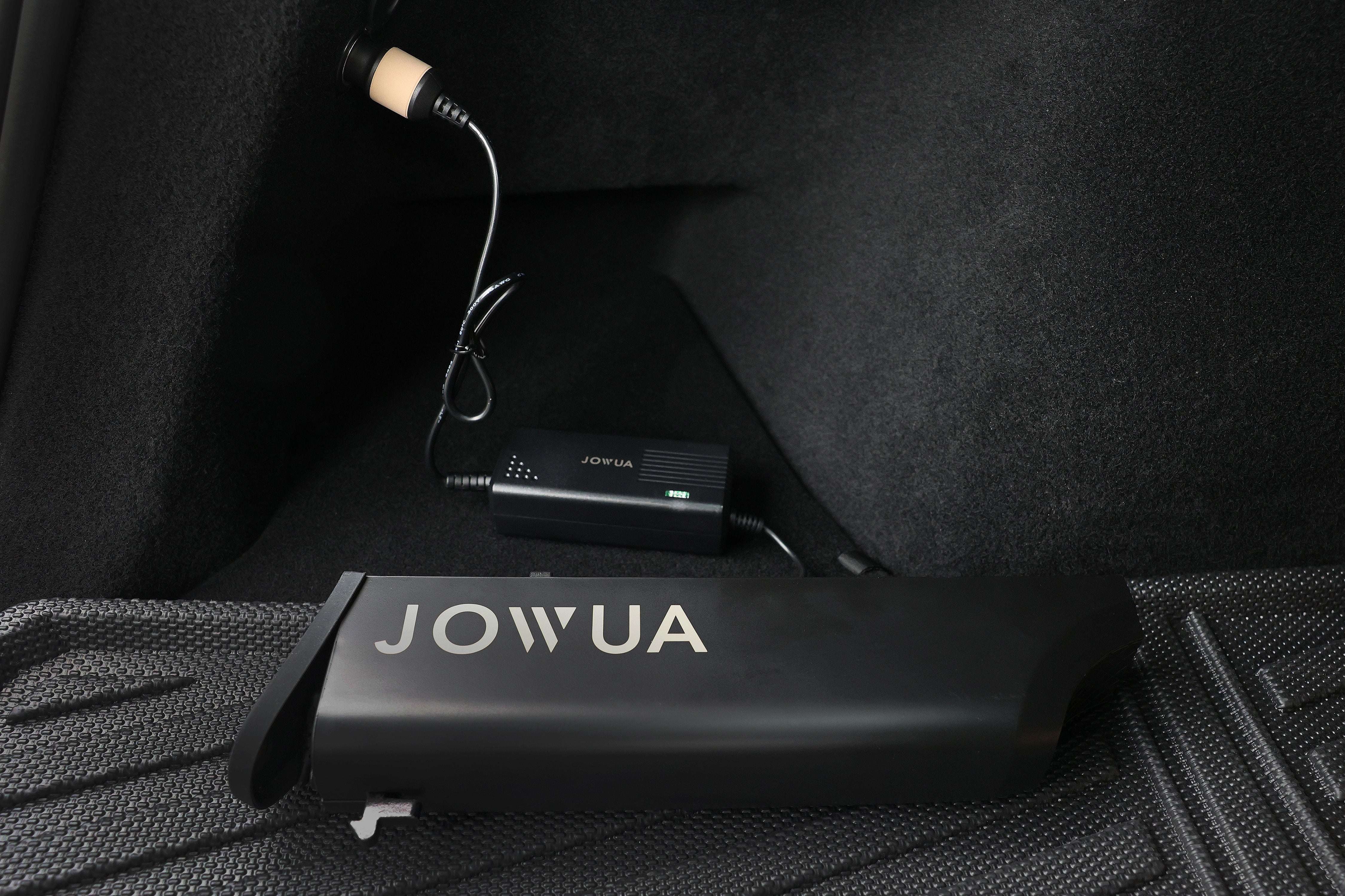 插上電源轉接器的黑色 JOWUA Taiwan 折疊電動滑板車|LR 放在汽車的黑色地板墊上,暗示對大容量電池的支持;背景是汽車的部分紋理內裝。.