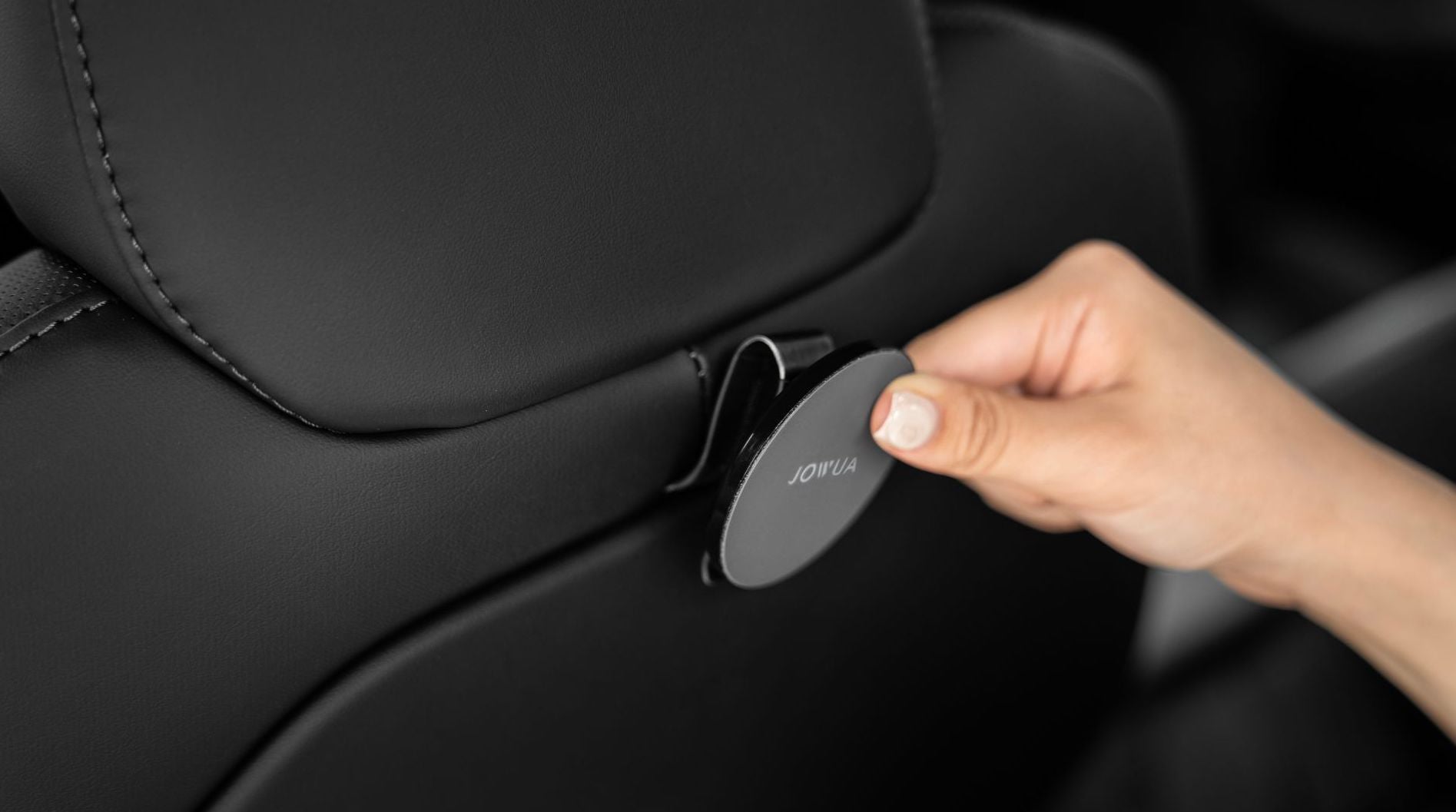 一手將圓形黑色 JOWUA 台灣 MagSafe 磁吸掛鉤組(型號 S/X/Y 煥新版)固定在 Model Y 椅背頭枕上,與時尚的黑色皮革內飾形成鮮明對比。.