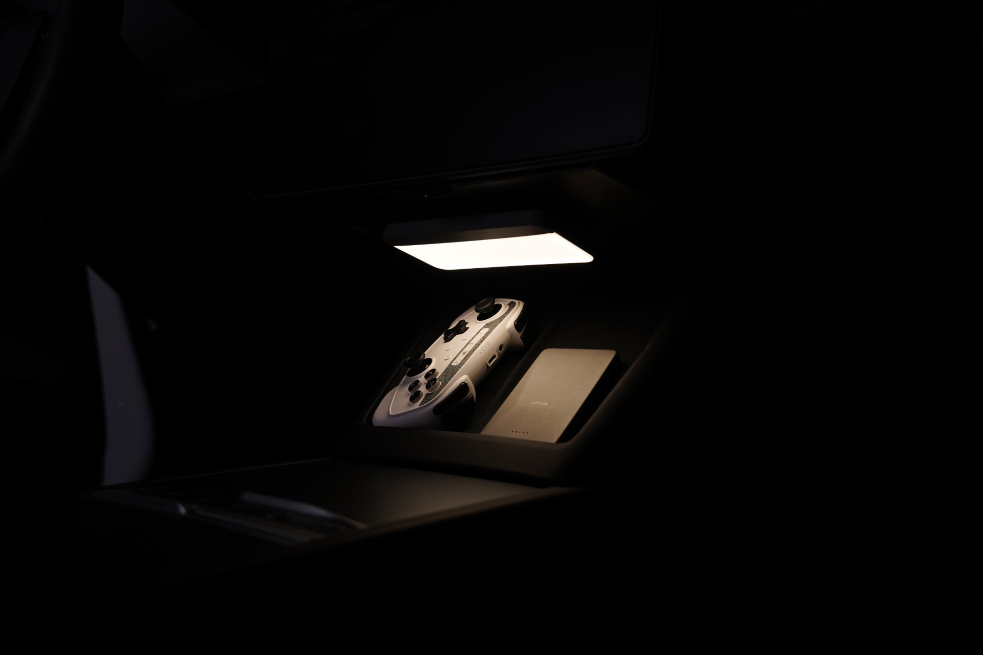 昏暗的車廂內,白色的遊戲控制器、閉合式的銀色筆記型電腦,以及 JOWUA Taiwan 的一幕瞭然【Model 3 煥新版|交車禮包】,都在一盞長方形頂燈的照耀下。.