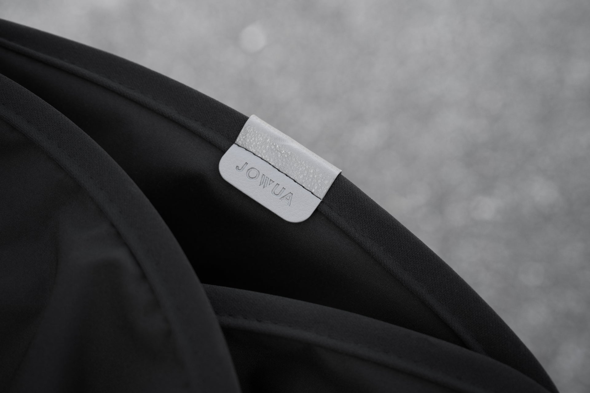 黑色織物特寫,上面印有“JOWUA Taiwan”的灰色標籤,體現了玻璃車頂遮陽簾 (Model S) 的時尚美感。背景呈現失焦的、有斑點的灰色表面。.