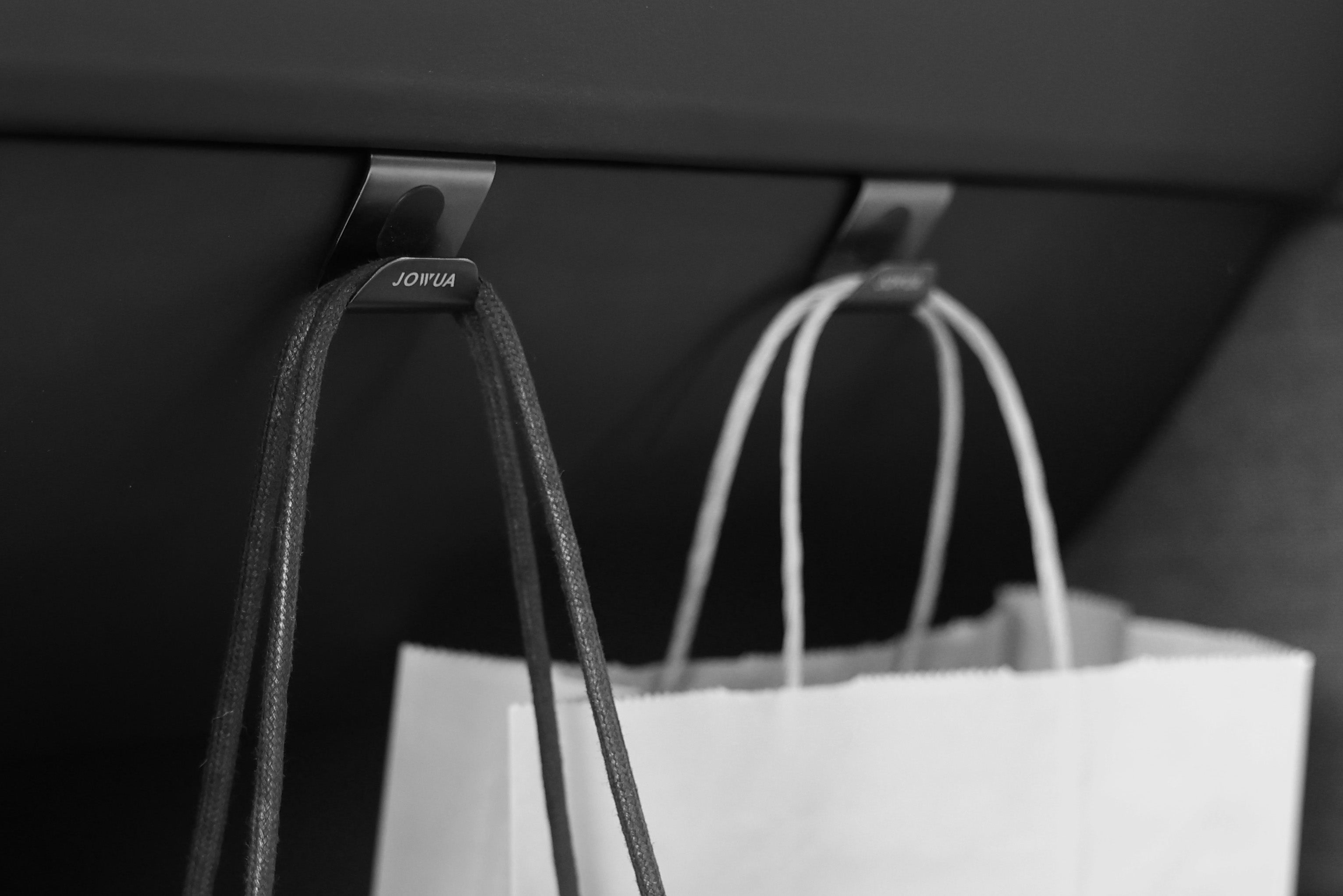 特寫圖顯示,架子下有兩個金屬掛鉤,時尚地掛著一個皮包和一個紙質購物袋。掛鉤上清晰可見 JOWUA Taiwan 品牌,體現了 Model 3 配件的時尚設計。場景以黑白色呈現,散發出永恆的精緻。.