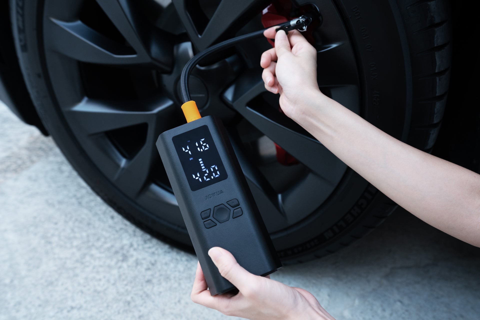 一個人使用具有高效車充功能的 JOWUA 台灣打氣機充電組合來為汽車輪胎充氣——一手拿著帶有數位顯示器的設備,另一隻手將噴嘴連接到閥門上。.