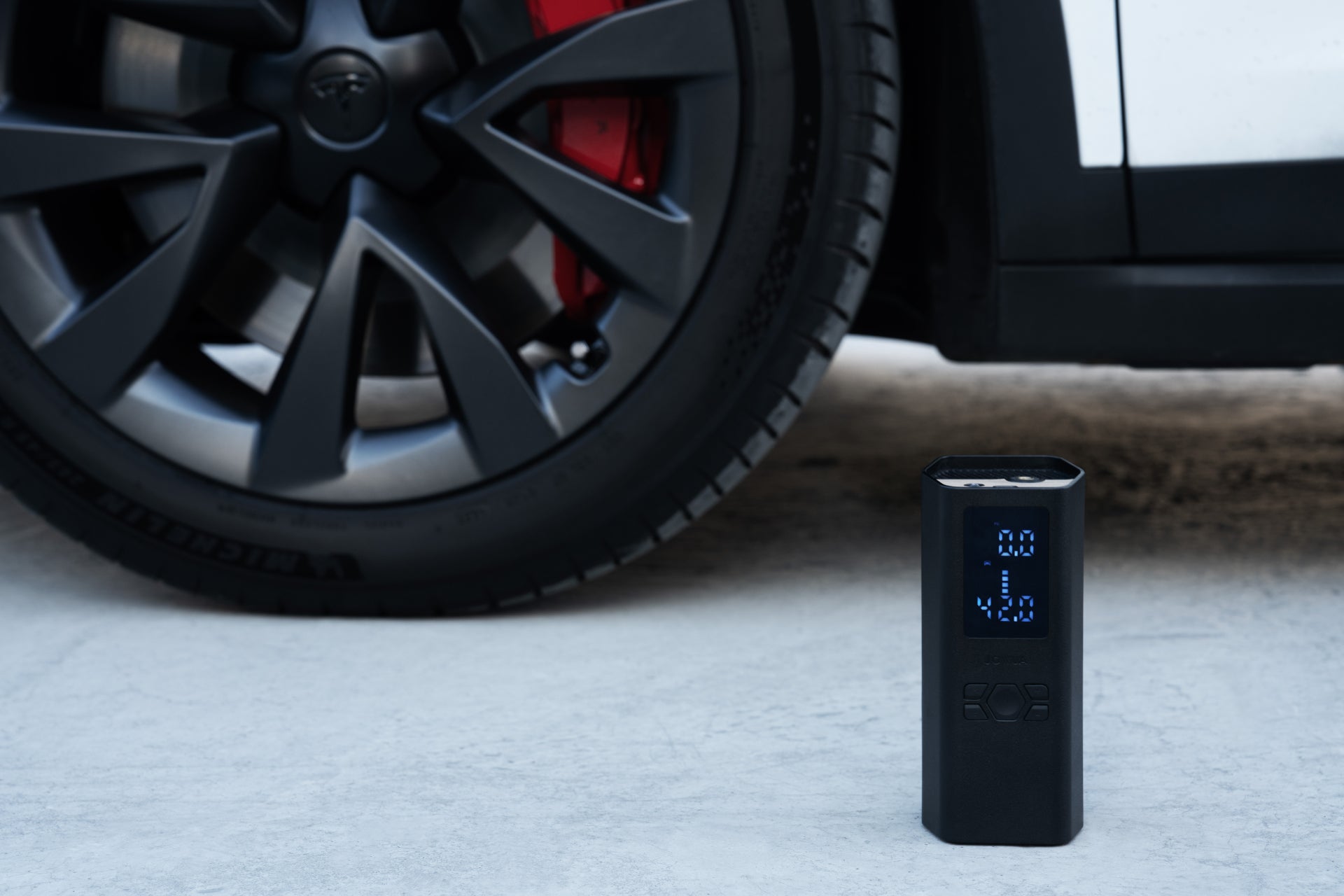 一台JOWUA台灣雙快充打氣機+氣嘴蓋組合,配有數位顯示器,放置在帶有紅色煞車卡鉗的汽車黑色車輪旁的地面上。汽車的白色外觀部分可見。.