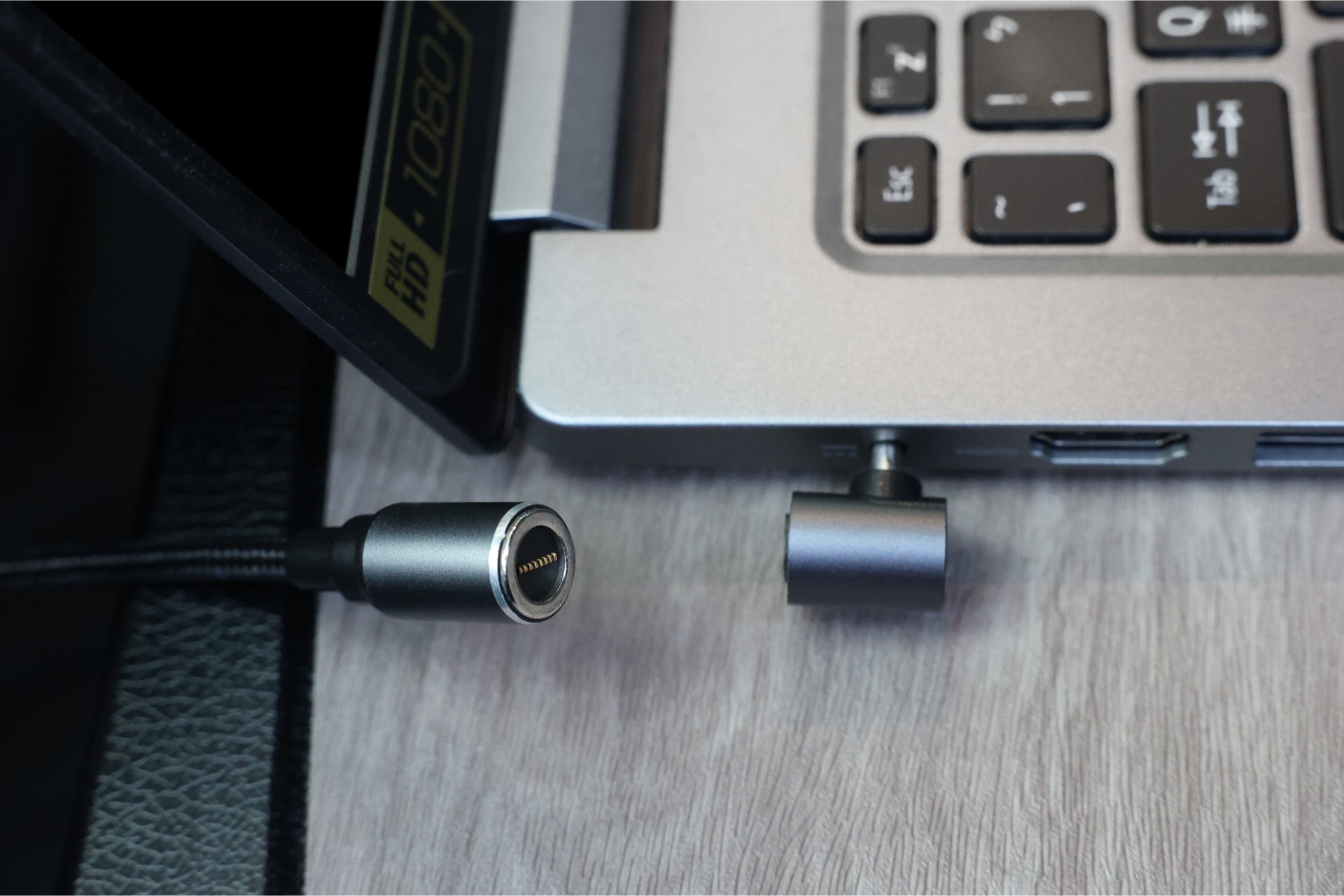 特寫顯示了筆記型電腦側面及其端口,以及 JOWUA Taiwan 100W 車充,其配備通用筆電車用磁吸充電線(專為非 USB-C 筆記型電腦設計),與端口分離。裝置放置在灰色木質表面上,部分鍵盤可見。.