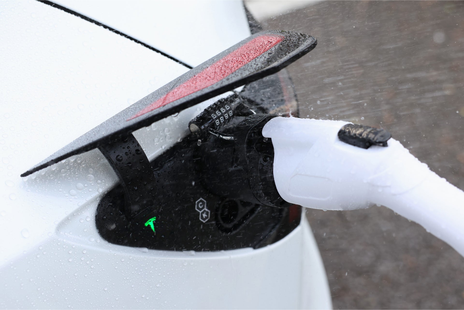 一輛白色電動車在雨中使用 JOWUA 台灣 J1772 轉 CCS2-AC 轉接器 (V3.5) 充電,展示其開放式充電埠。白色電纜連接,水滴增強車輛的韌性和先進的溫度保護晶片技術。.