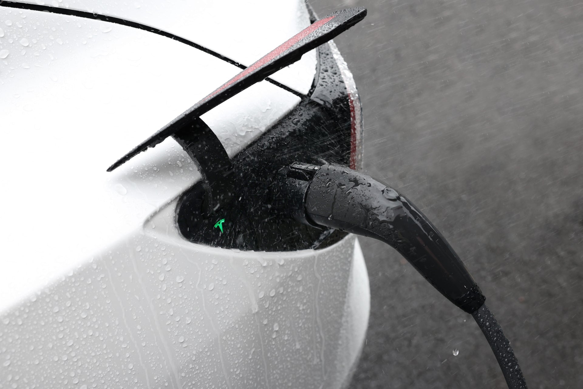 當水滴閃閃發光時,JOWUA 台灣 J1772 至 TPC(NACS) 轉接頭為雨中的電動車提供動力。充電線連接到白色車輛的後部,透過打開的連接埠可以看到發光的箭頭指示器。.