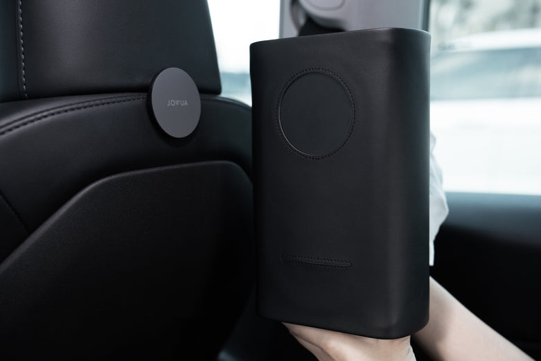 一個人拿著 JOWUA 台灣 MagSafe 磁吸掛鉤(型號 3 2018-2024/型號 Y 2021-2024),這是一款與 iPhone MagSafe 兼容的時尚黑色設備,旁邊是一個汽車座椅,配有圓形 JOWUA 附件,用於優化車內收納空間。.