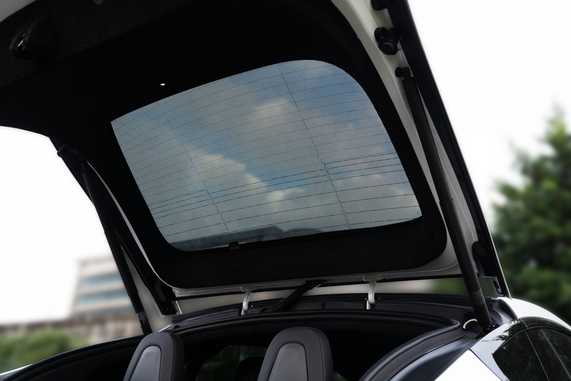 圖片顯示了 Model X 2021+ 的內部打開的後行李箱,透過後窗可以看到多雲的天空、綠色植物和一棟建築物。內部設有兩個頭枕,並配有 JOWUA 台灣尾門+三角窗遮陽簾,可遮蔽紫外線,增加舒適度。.