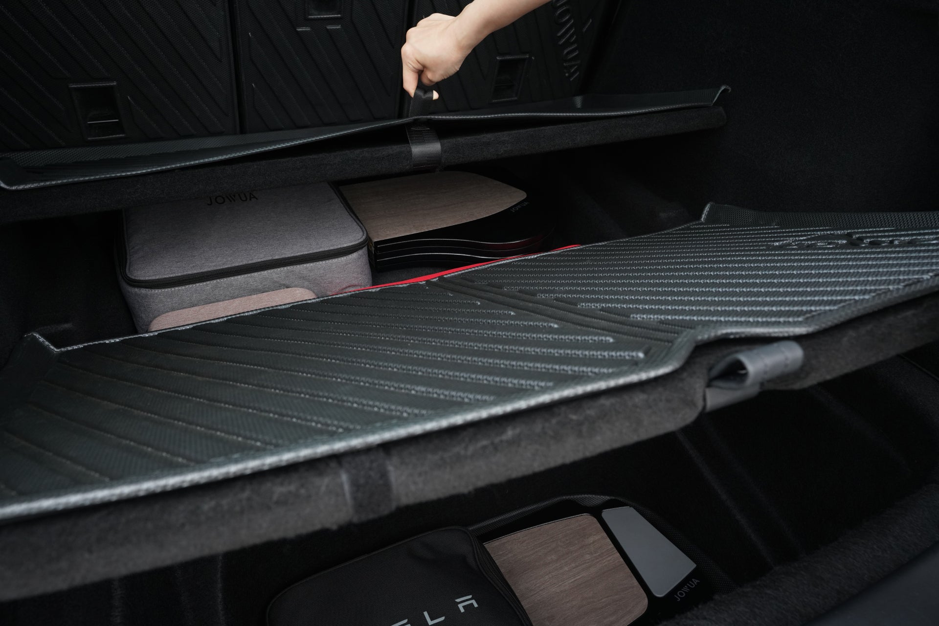 一手抬起特斯拉 Model Y 後行李箱的貨蓋,露出整齊的隔間和 Tesla 品牌包袋下方的 JOWUA 台灣易開式防水行李廂墊(椅背墊可加選,2025+/2021-2024 適用)。.