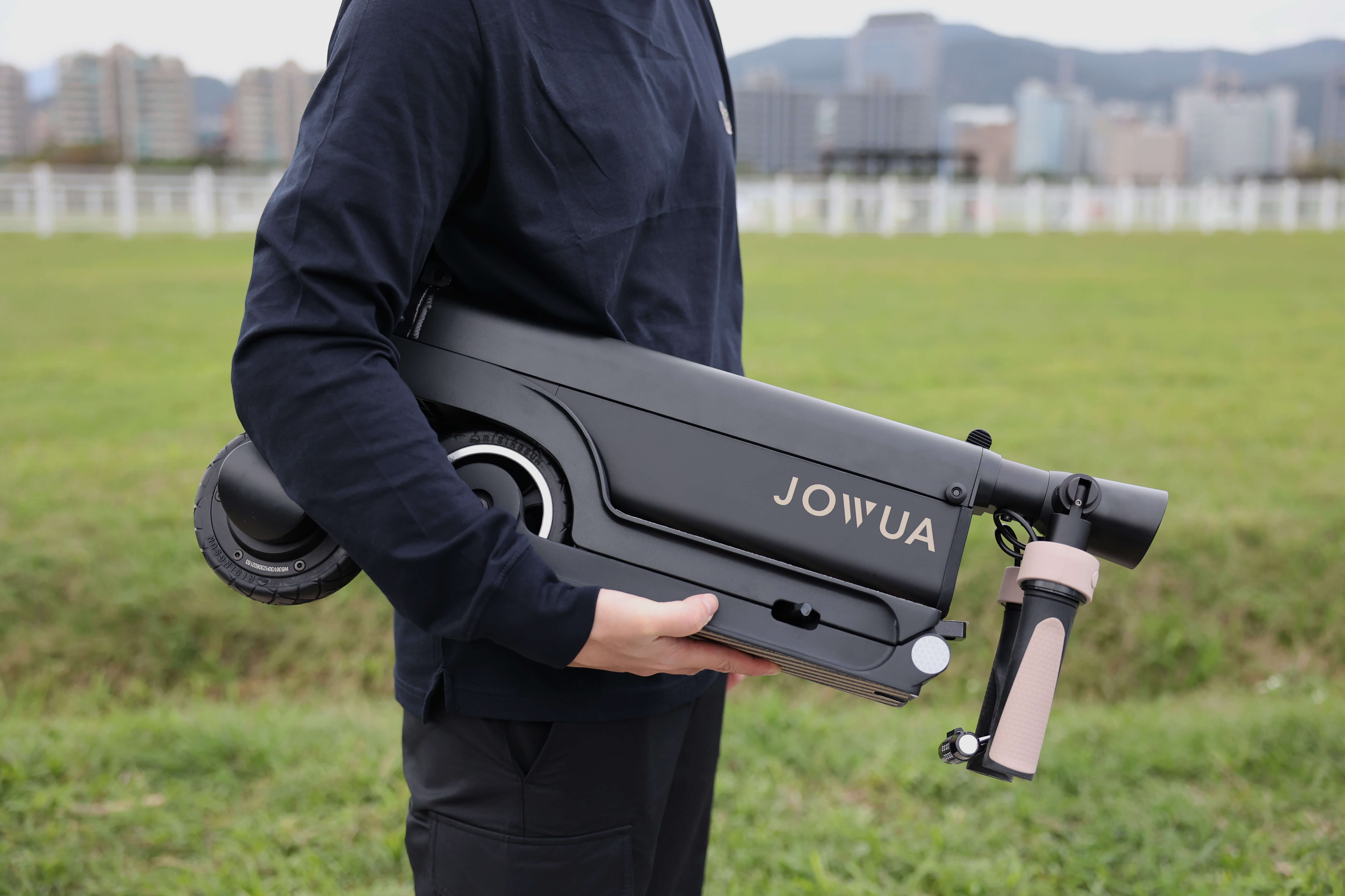 一位穿著深色衣服的人,手持折疊的 JOWUA Taiwan 折疊電動滑板車|LR,配備大容量電池,站在草地上,背景是城市景觀和山脈。.