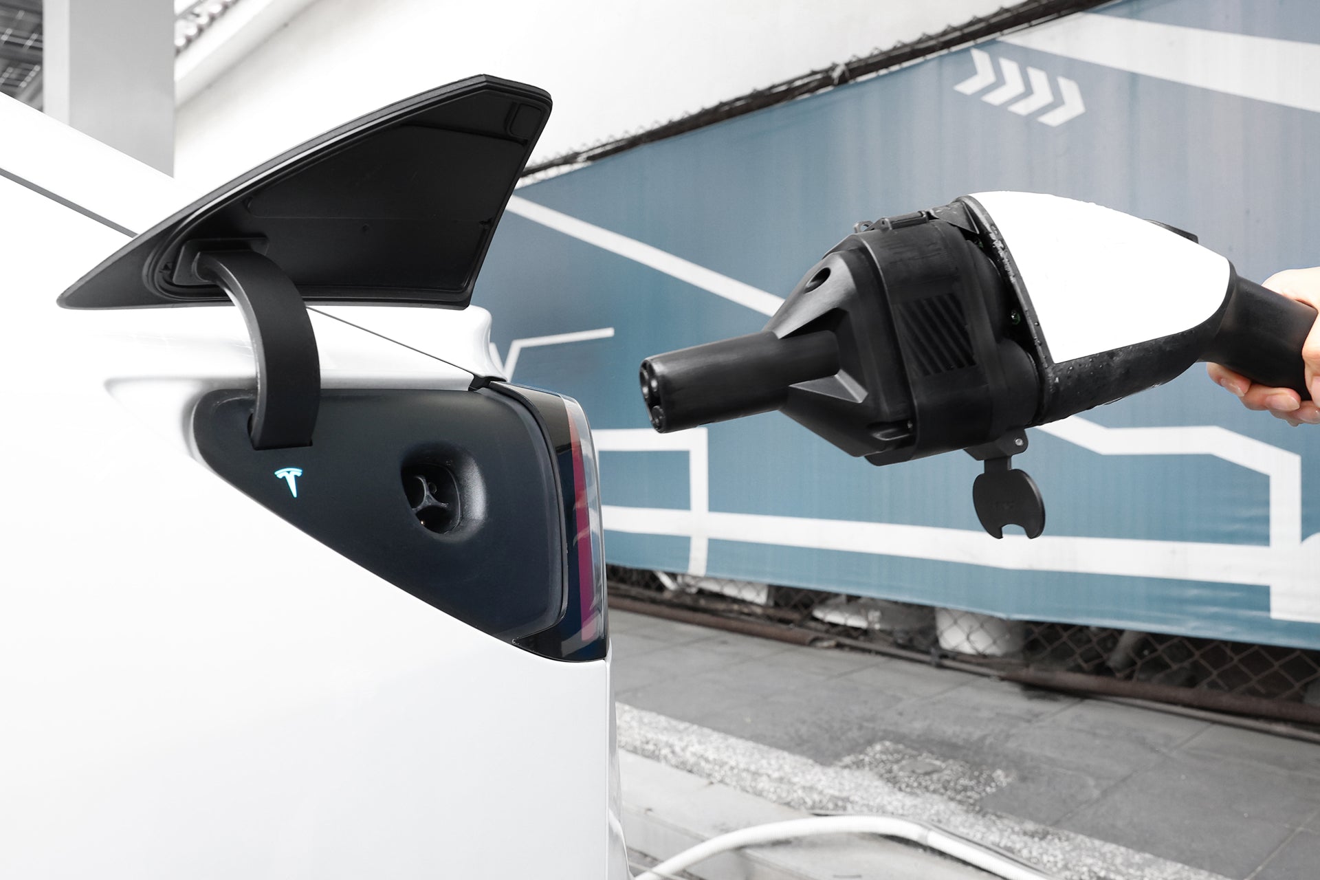 一個人拿著 JOWUA Taiwan CCS1 to NACS (TPC) DC & AC 快慢充轉接頭轉接器靠近一輛白色 Tesla 的充電口,準備連接。背景是一面藍白幾何牆。.