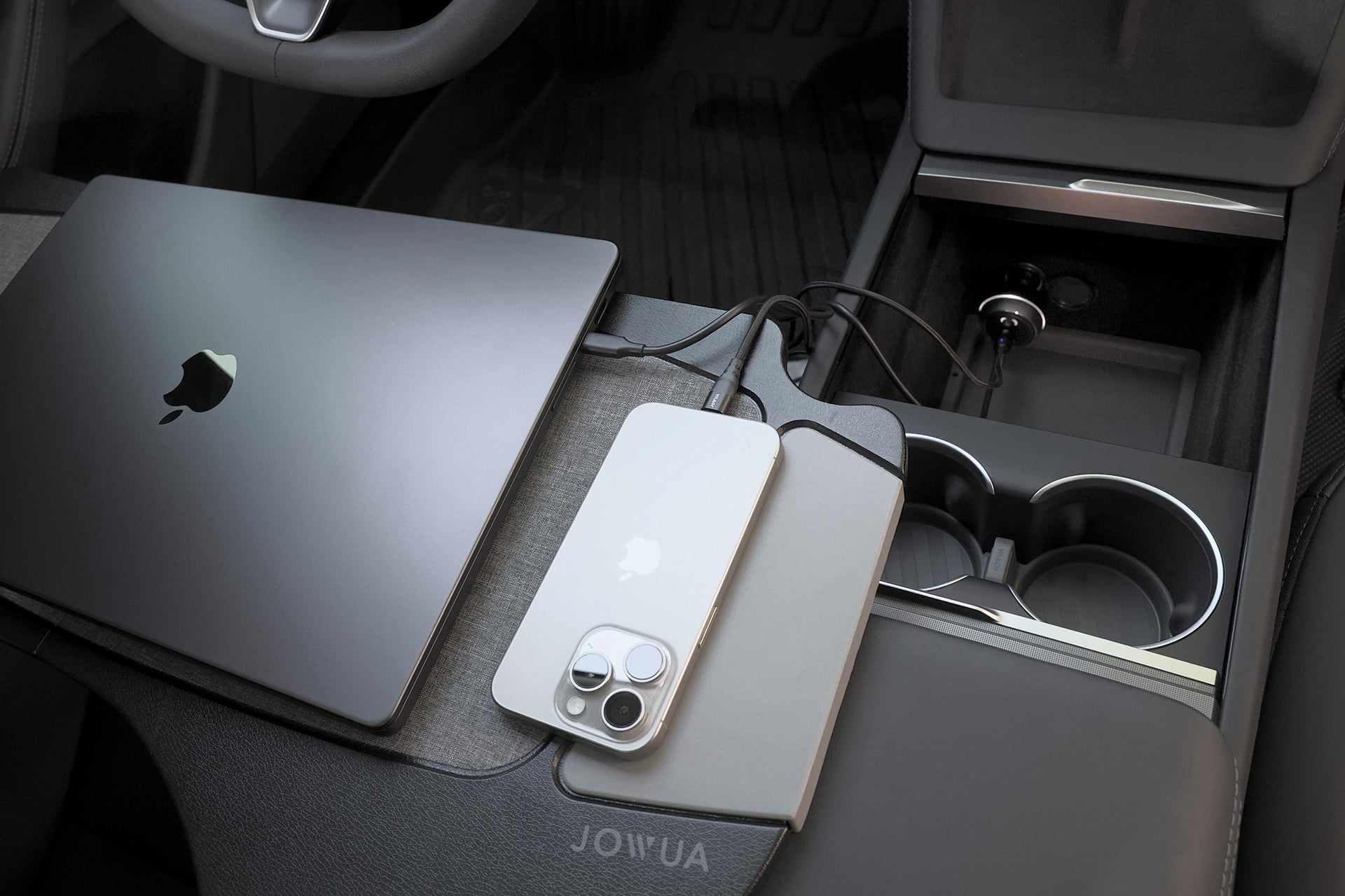 MacBook 和 iPhone 正在中控台上使用 JOWUA 台灣 165W PD3.1 高效能車充三埠輸出連接電纜進行充電。設備旁邊還可以看到杯架和小隔間。.