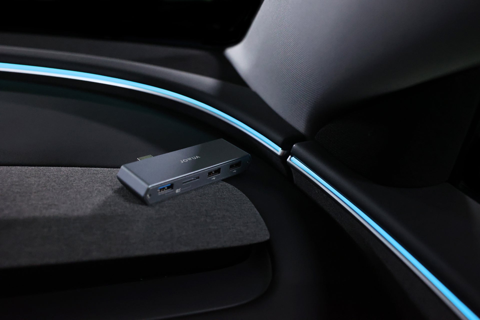 JOWUA Taiwan Tesla 雙用儲存 HUB (讀卡器 & 原廠 USB) 置於 Tesla 車內深色紋理表面上,並由車內面板上的藍色環境照明照亮。.