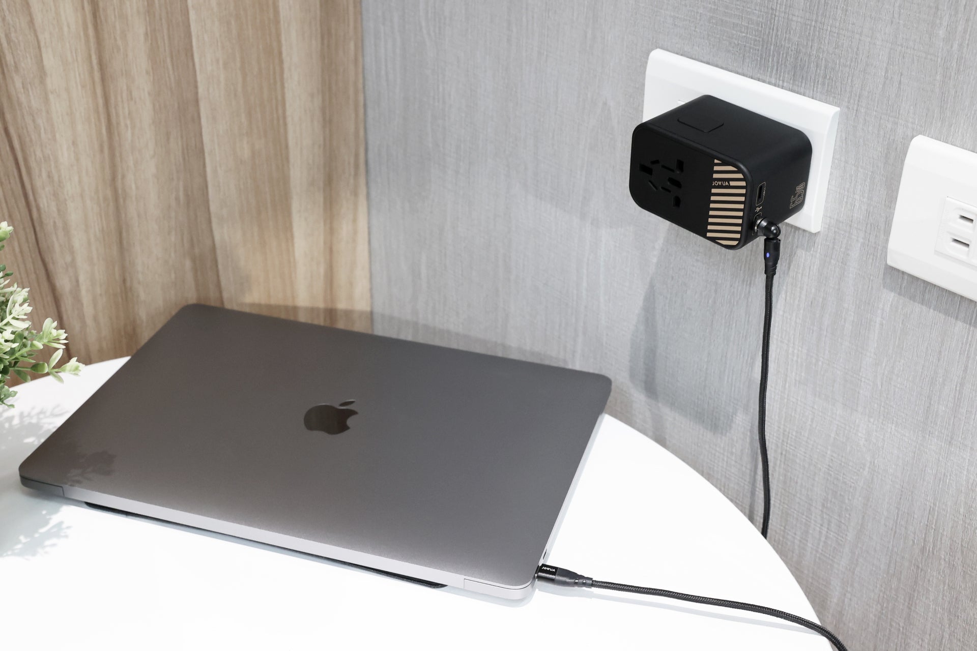 一台印有 Apple 標誌的灰色筆記型電腦放在一張白色圓桌上,插入一個 JOWUA Taiwan 65W 旅行萬國插頭氮化鎵充電器。黑色電源轉接器連接到牆上的插座,可提供快速充電,並凸顯中性色調牆壁上的一株小植物。.