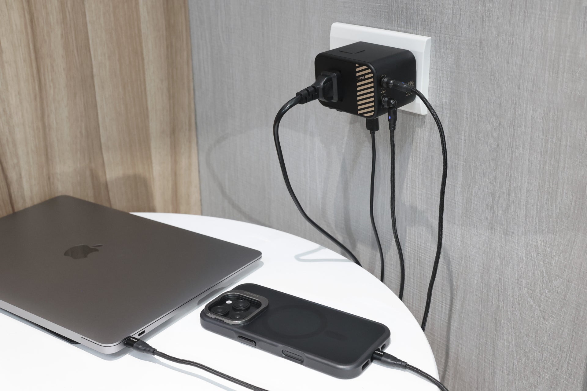 一台筆記型電腦和智慧型手機放在一張白色的桌子上充電,微微反光,連接到牆上插座的 JOWUA Taiwan 65W 旅行萬國插頭氮化鎵充電器。此轉接器支援多條電纜,可高效使用和快速充電。.