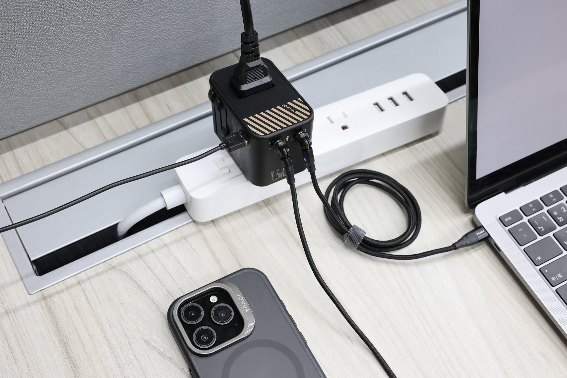 一部智慧型手機、一台筆記型電腦和一台帶有電纜的JOWUA Taiwan 65W旅行萬國插頭氮化鎵充電器整齊地擺放在一張淺色木桌上。此充電器具有多重安全保護功能,可連接到電源板,以便在裝置附近快速充電。.