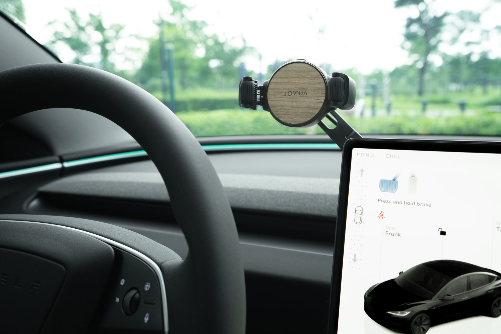 該車內裝配備方向盤、適用於 Model 3/Y 和 Cybertruck 的「JOWUA Taiwan」Qi2 無線充電儀錶板手機支架,以及帶有汽車示意圖的觸控螢幕顯示器。透過窗戶,可以看到鬱鬱蔥蔥的樹木和綠地。.