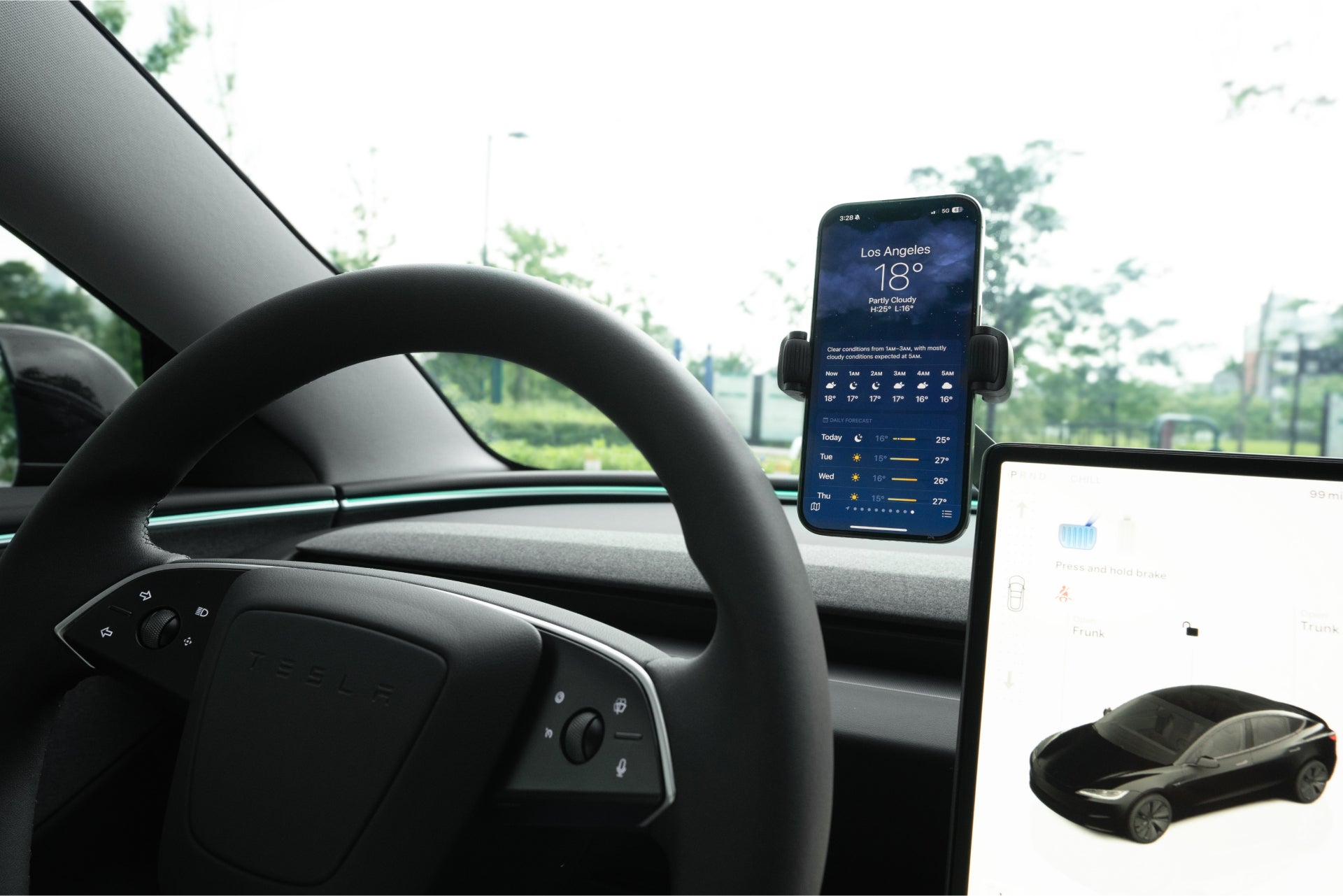 車內包括方向盤和台灣 JOWUA 的六維車架 Qi2_滾輪款,可以以 15W 的功率為智慧型手機無線充電,並提供洛杉磯天氣更新。資訊娛樂螢幕上顯示了一輛汽車的插圖,擋風玻璃視圖顯示了陰天下的茂盛綠色植物。.
