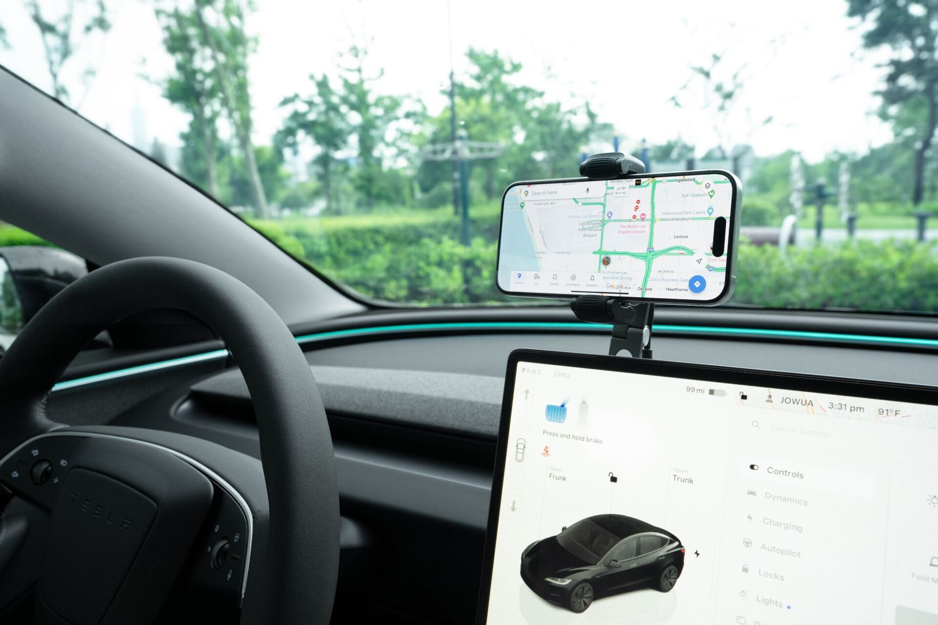 帶有方向盤的汽車內裝、顯示汽車示意圖的儀表板螢幕以及儀表板上的智慧型手機,採用 JOWUA 台灣六維車隱藏架 Qi2_滾輪款,可實現 15W Qi2 無線充電。透過擋風玻璃,可以看到前方道路兩旁鬱鬱蔥蔥的綠樹。.