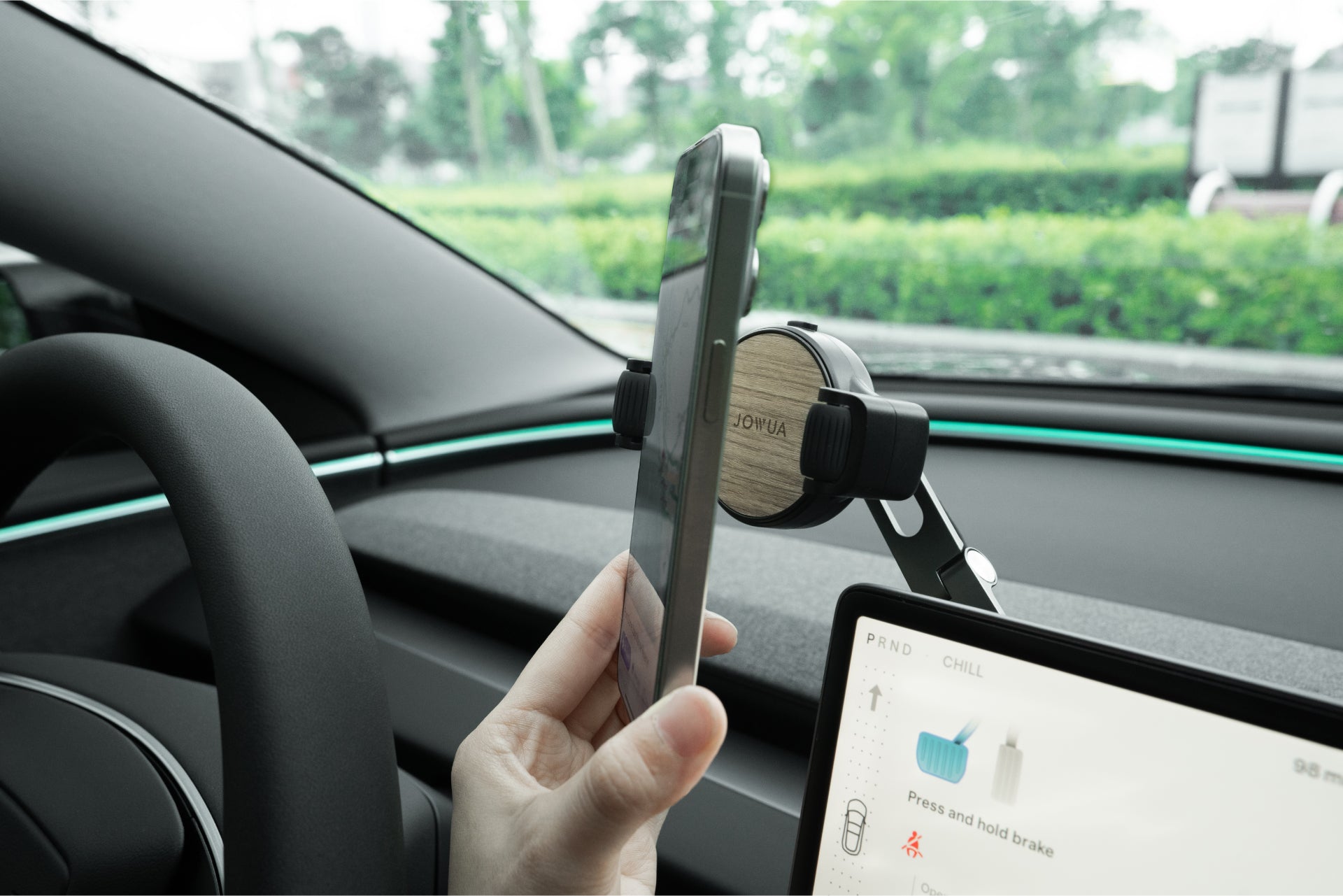 有人正在將智慧型手機放入JOWUA台灣六維車架Qi2_滾輪款(Model 3/Y煥新版、Cybertruck專用)車內。數位顯示器顯示駕駛數據,同時可以透過擋風玻璃看到綠色植物。.