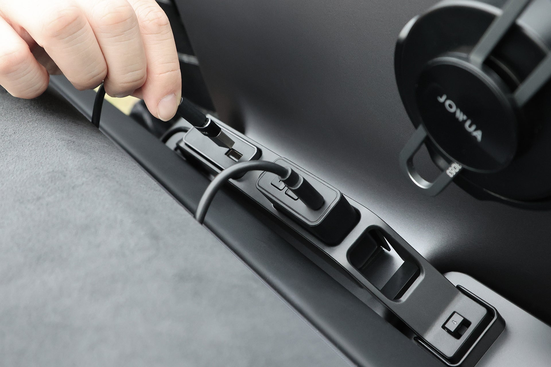 一隻手將充電電纜穿過一幕瞭然【Model Y 煥新版|交車禮包】電纜收納架,由台灣 JOWUA 製造,置於 Model Y 的儀表板上,附近可見 JOWUA 的手機支架。.