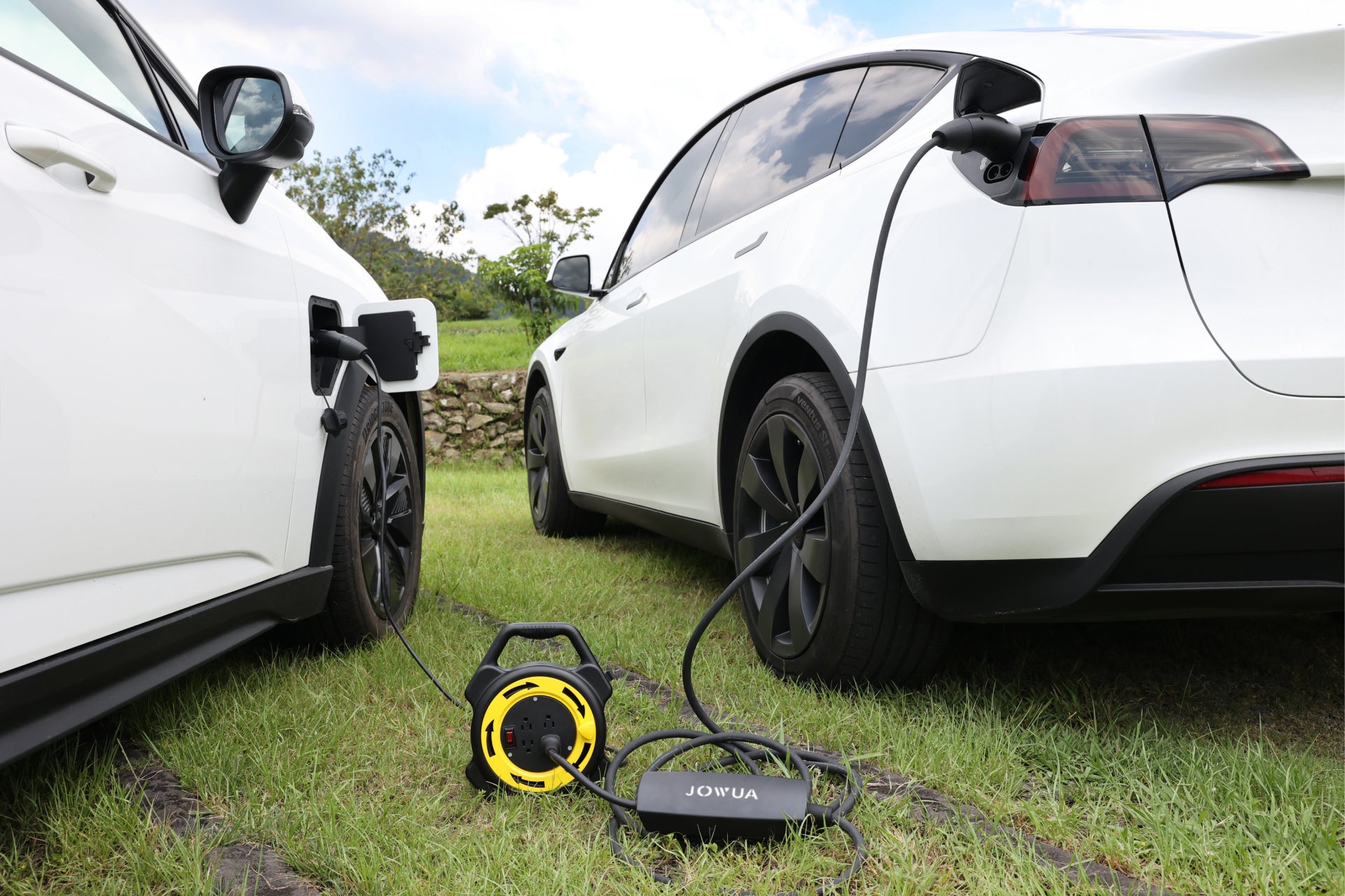 草地上停放的兩輛白色電動車透過充電線連接。一條電纜連接到地面上的 JOWUA Taiwan 可伸縮式V2L放電插座 J1772 設備,作為 V2L能源轉換系統。背景為多雲的天空下,石牆和綠色植物矗立著。.
