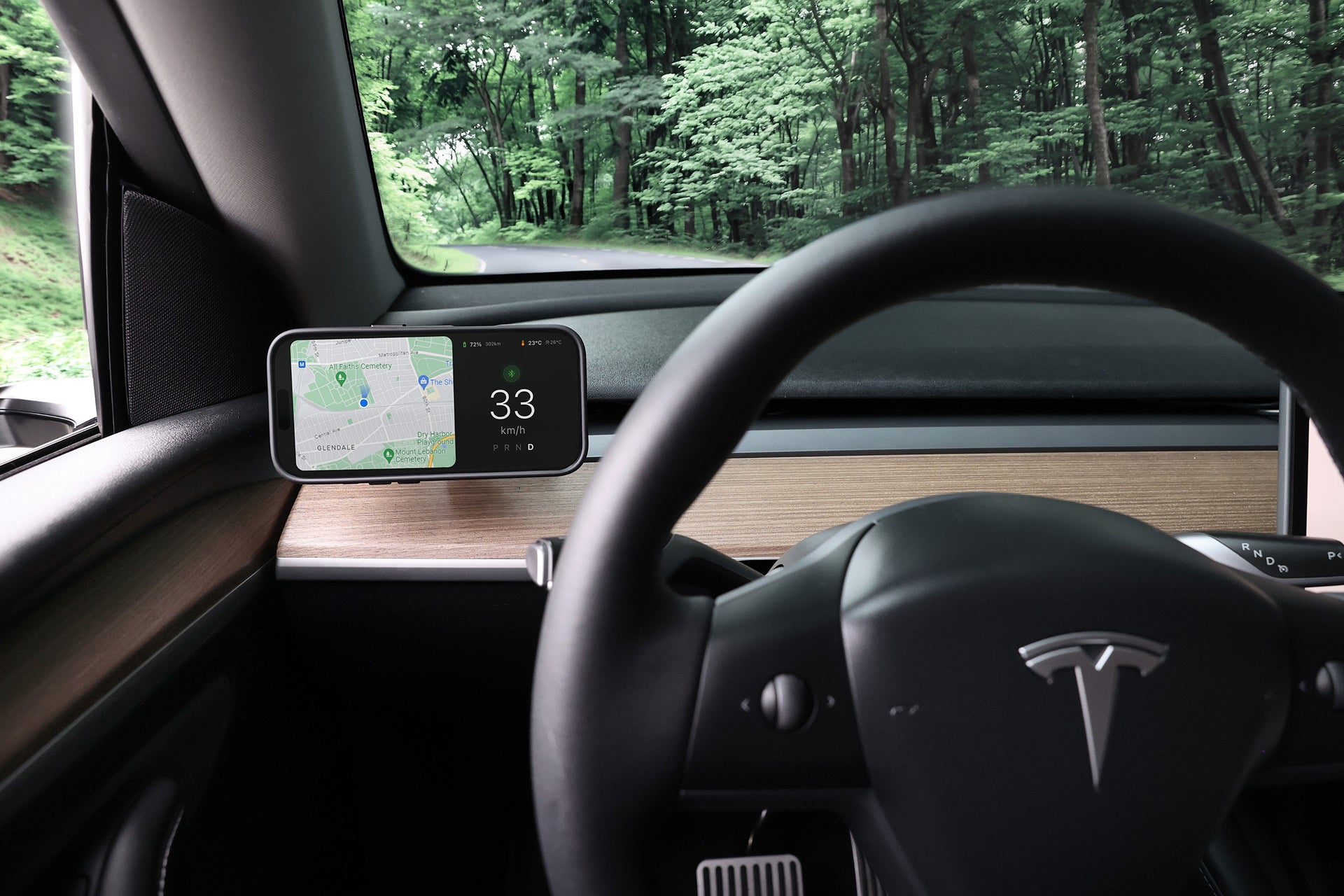 從 Tesla Model 3/Y (2018-2024) 車內觀看,出風口設有 JOWUA Taiwan Qi2 製冷出風口 MagSafe 車架,可啟用 MagSafe 無線充電,同時在風景秀麗的森林道路上行駛,特色是方向盤和數位顯示器。.