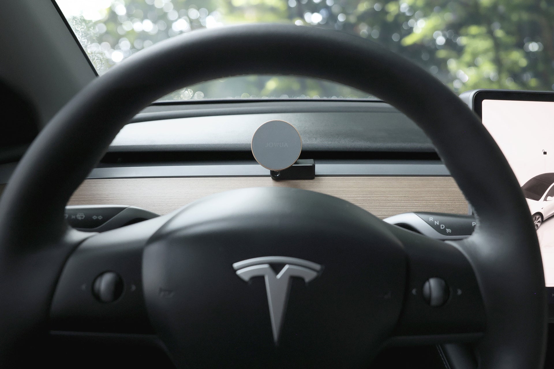 從 Tesla 車輪後方觀看,顯示 Tesla 標誌、木製儀表板、整合 MagSafe 的 JOWUA Taiwan Qi2 製冷出風口 MagSafe 車架 (Model 3/Y 2018-2024)。樹木透過擋風玻璃出現。.