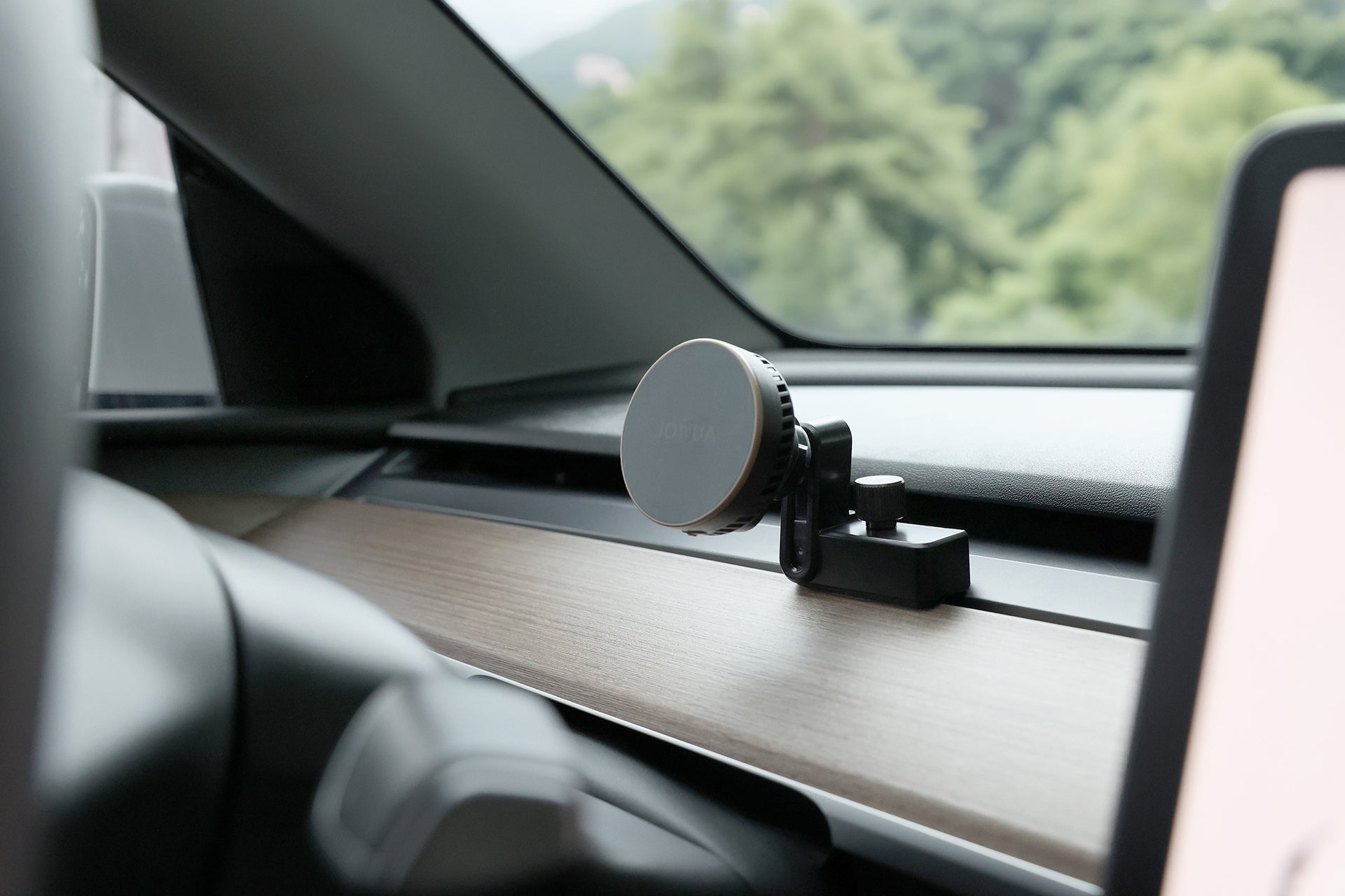 一輛 JOWUA Taiwan Qi2 製冷出風口 MagSafe 車架 (Model 3/Y 2018-2024) 裝在汽車出風口上,背景是透過擋風玻璃模糊的綠樹。.