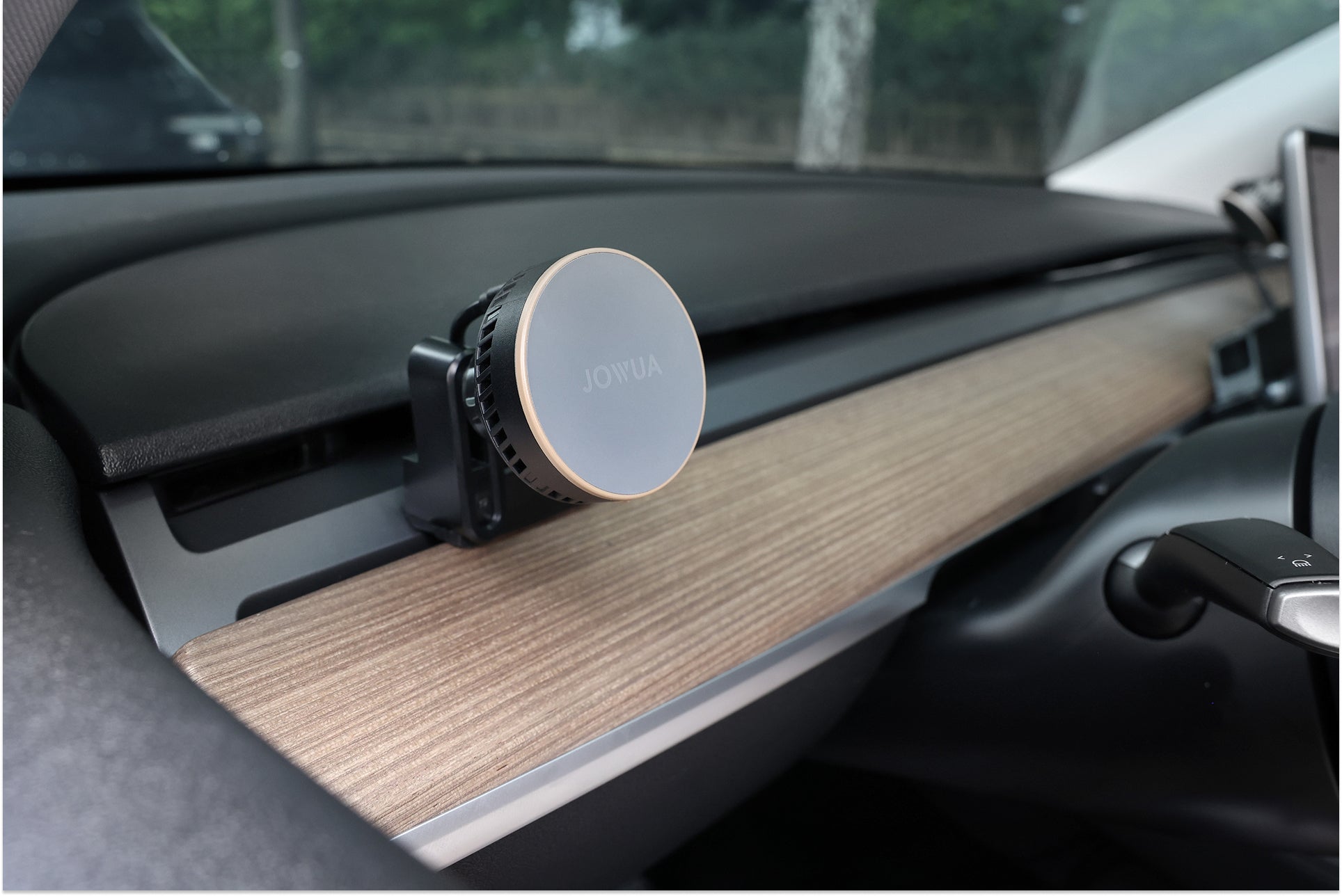 一輛 JOWUA Taiwan Qi2 製冷出風口 MagSafe 車架 (Model 3/Y 2018-2024) 夾在木質紋理儀表板上方的汽車出風口上,部分方向盤和車窗也清晰可見。.