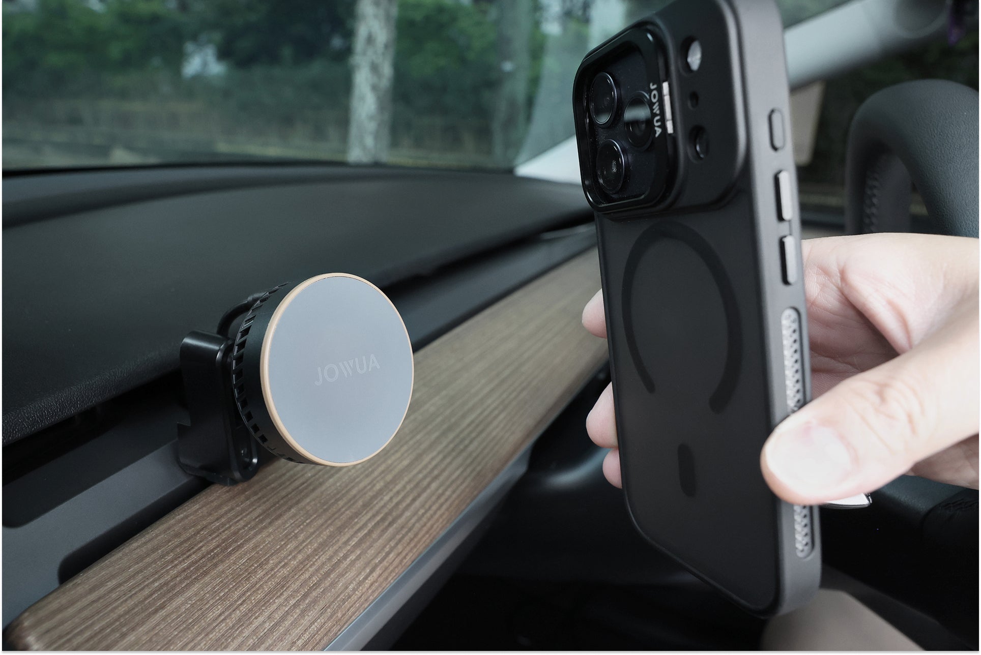 一隻手在木飾儀表板上的 JOWUA Taiwan Qi2 製冷出風口MagSafe 車架 (Model 3/Y 2018-2024)附近拿著一個裝有 MagSafe 保護套的智慧型手機,從擋風玻璃可以看到樹木和籬笆。.