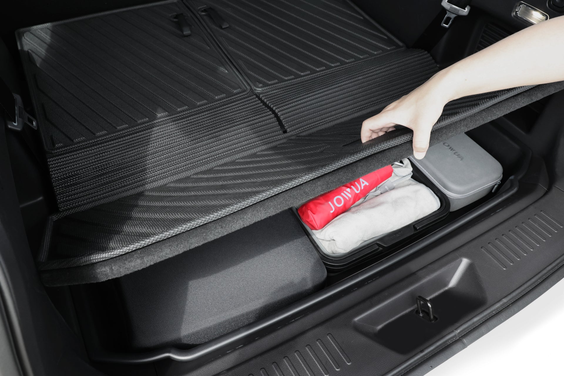 一隻手掀開汽車後備箱裡的 JOWUA Taiwan 立體防水行李廂墊+椅背墊組合 (LUXGEN n⁷ 7人座),露出一個儲物格,裡面有紅色水壺、折好的白色毛巾和灰色箱子。.