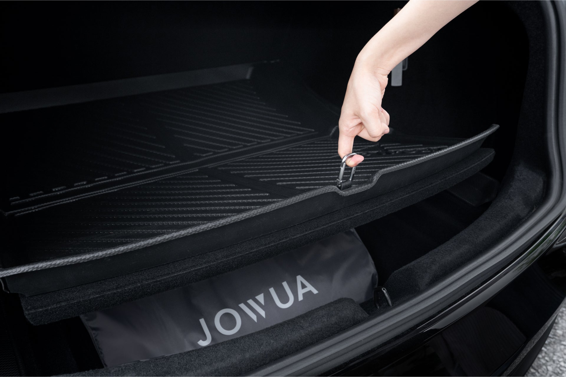 一隻手掀開 Tesla Model 3 後備箱內的黑色貨墊,露出下方存放的 JOWUA Taiwan 電墊專案【Model 3 煥新版|交車禮包】。.