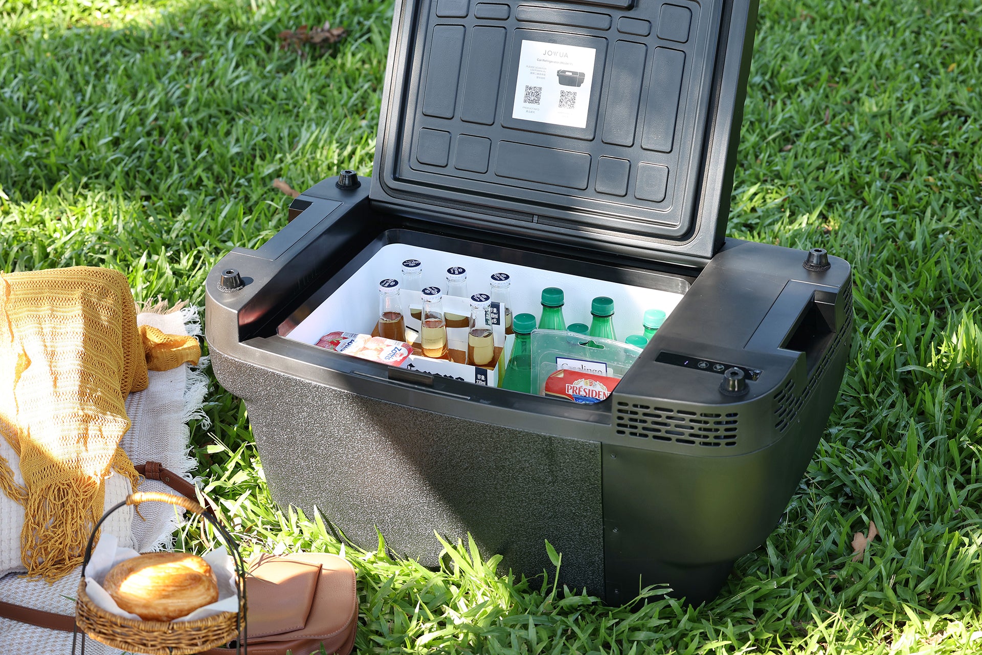 一台黑色 JOWUA 台灣製 Type-C & 點菸器 雙軌車用行動冰箱(Model Y 全車系)敞開著置於草地上,裡面裝滿了瓶裝飲料和零食。 旁邊的野餐區鋪著黃色野餐墊,擺放著籃子和食物——這正是戶外放鬆的完美組合,搭配 Type-C 穩定供電,確保您的飲品隨時冰涼。.