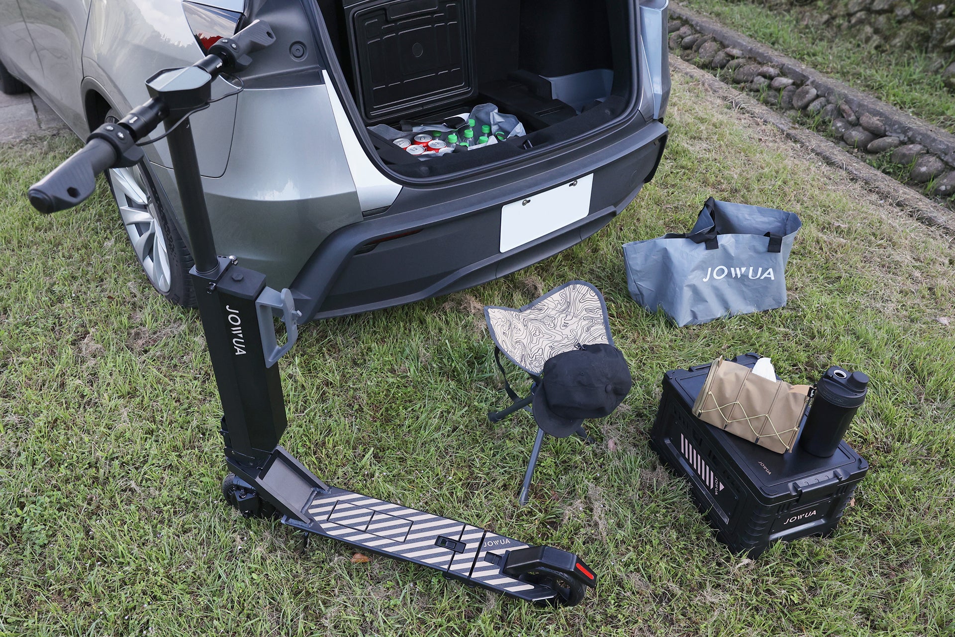 摺疊式電動代步車、攜帶式座椅、JOWUA Taiwan Type-C&點菸器雙軌車用行動冰箱 (Model Y全車系) 35L、收納箱、袋子和熱水壺,排列在開放式汽車後備箱後方的草地上,供野餐或戶外活動使用。.