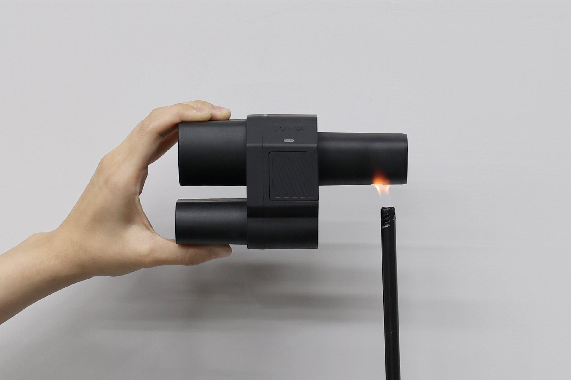 一隻手中,一個光滑的圓柱形黑色裝置用它的火焰點燃了一根黑色蠟燭。這種現代的精確度讓人想起了 JOWUA 台灣的 CCS2 到 NACS (TPC) 直流轉接頭。背景是一面普通的灰色牆壁。.
