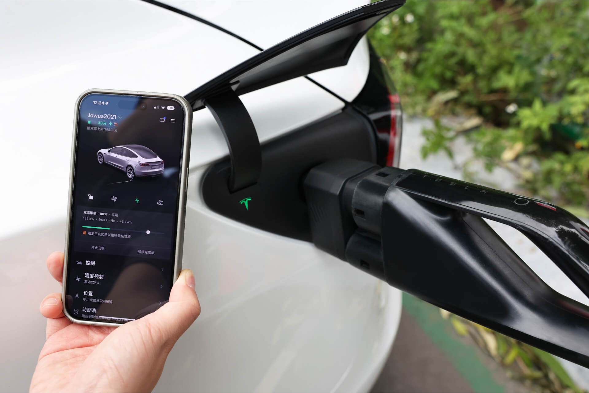 某人拿著一部顯示汽車詳細資訊的智慧型手機,旁邊是一輛透過其發光的綠色連接埠充電的電動車,使用 JOWUA 台灣 CCS2 至 NACS (TPC) 直流轉接頭進行高效充電,背景中可以看到植物。.