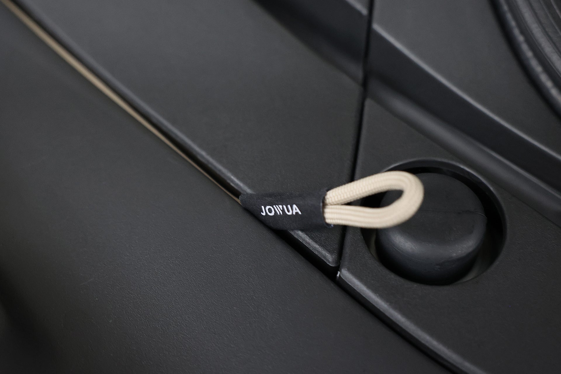 黑色表面上有一條米色的環形繩,上面標有“JOWUA Taiwan”,緊貼著小按鈕旁邊的凹槽。這個元素反映了納智捷LUXGEN n⁷前行李箱空間充電槍緊急釋放延長拉繩的詳細設計。.