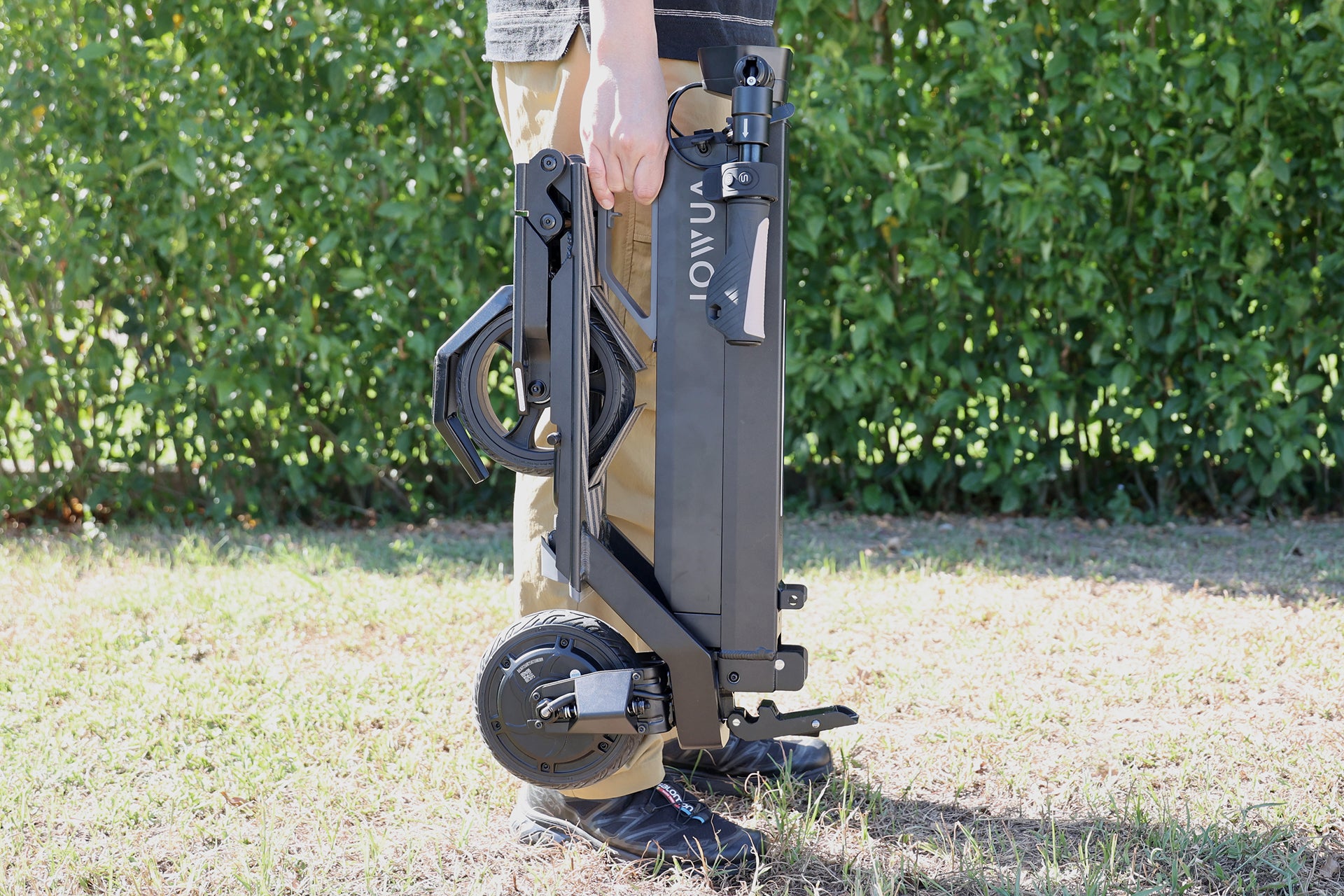 一個人站在草地上,手持折疊的 JOWUA Taiwan 折疊電動滑板車|演出畫框,背景為綠意。.