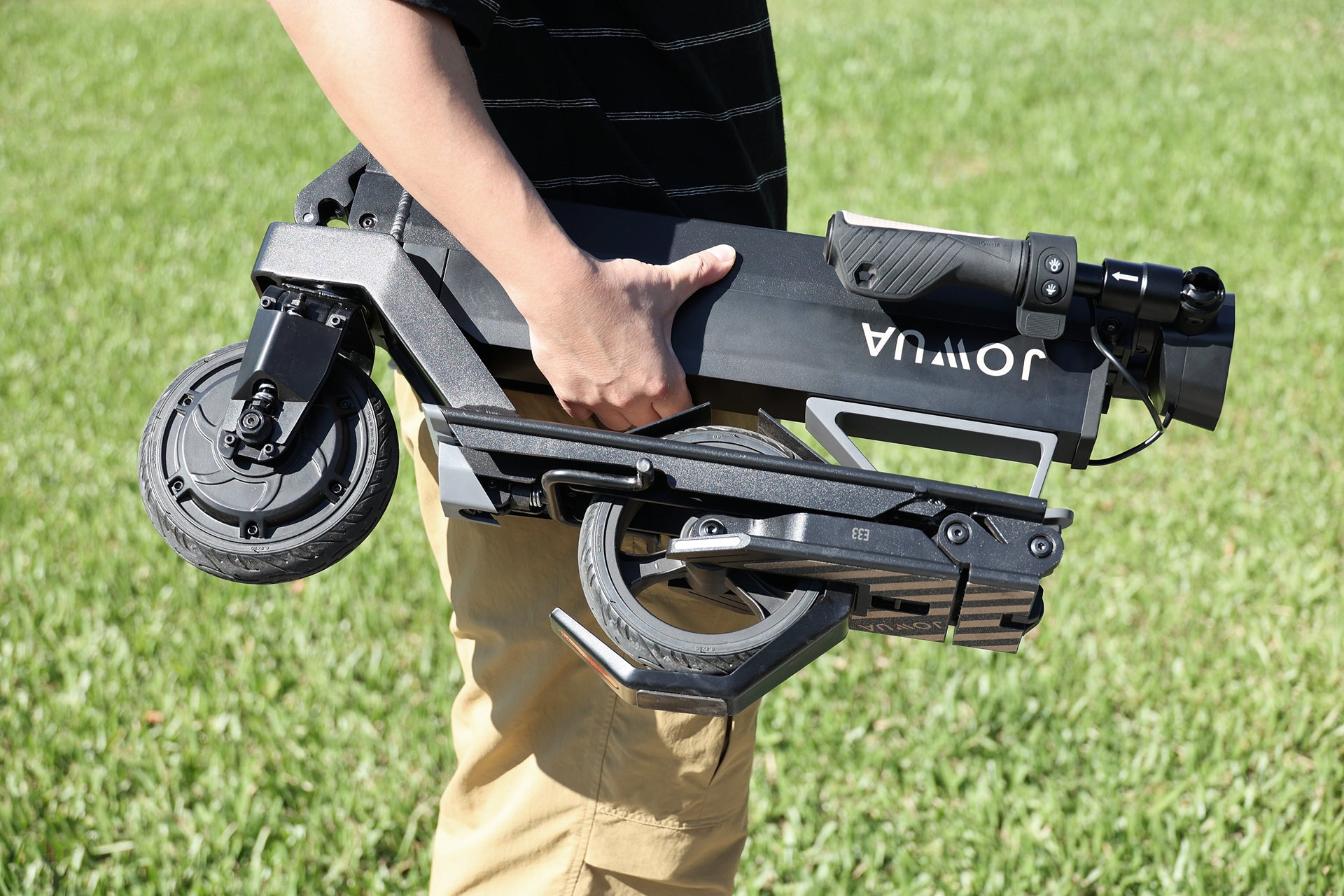 一個身穿棕褐色褲子和黑色襯衫的人站在綠色草地上,手持折疊的 JOWUA Taiwan 折疊電動滑板車|Performance with 電池容量升級。.