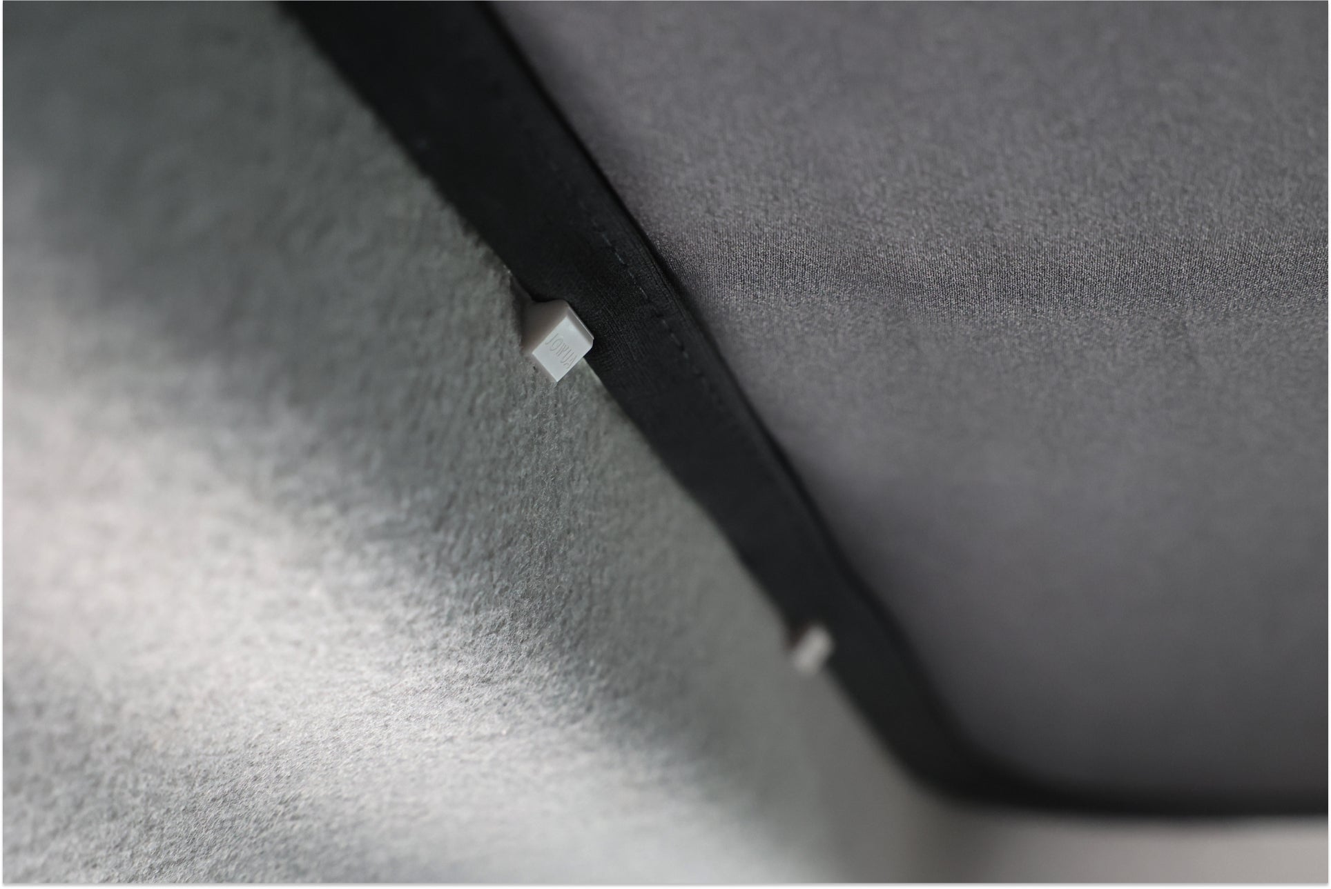 JOWUA Taiwan 玻璃車頂遮陽簾 (LUXGEN n⁷) 的紋理灰色織物詳細視圖,顯示出帶有兩個白色夾子的對角線接縫,類似於車頂遮陽簾中的接縫。柔和的燈光在材質上營造出柔和的陰影。.