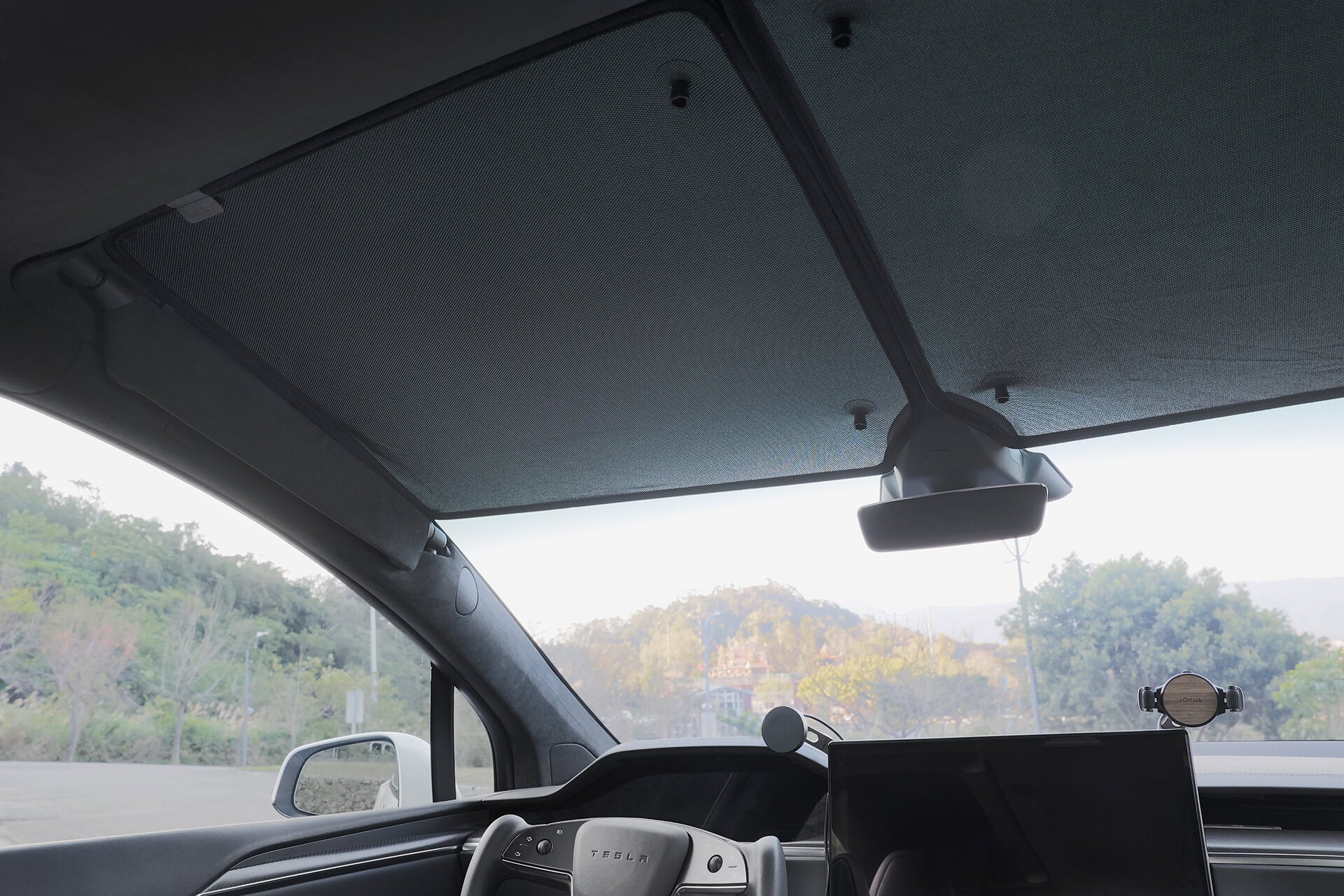 特斯拉車內景,儀表板、方向盤和 JOWUA Taiwan 分離式遮陽簾 (Model X) 覆蓋全景隔熱車頂。樹木和山丘透過擋風玻璃清晰可見,為每次駕駛增添舒適與風格。.