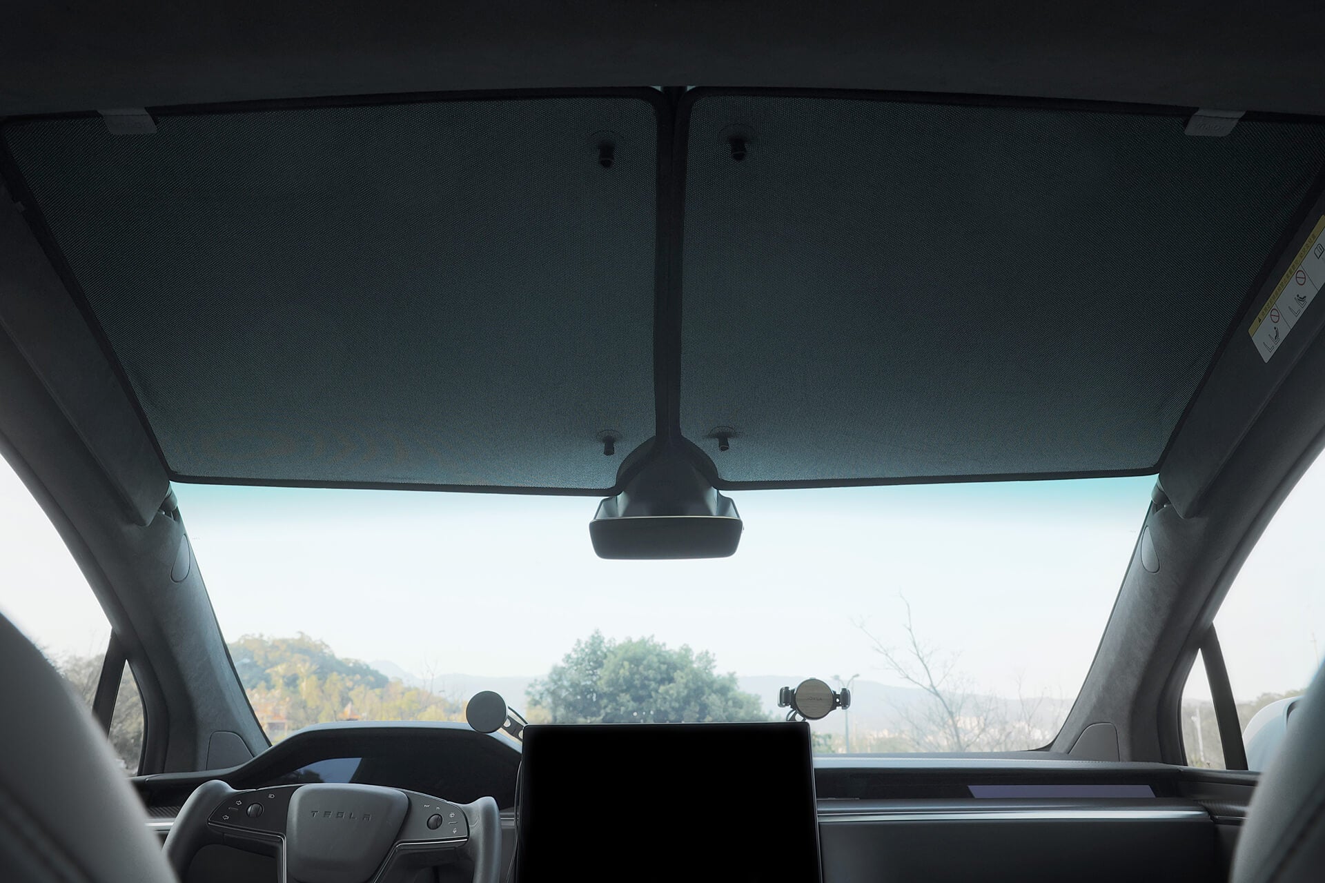 在 Model X 內部,可以看到擋風玻璃和部分內部裝飾,其中包括中央顯示器和後視鏡。從外觀上看,可以看到樹木上方晴朗的天空。 JOWUA 台灣分離式遮光簾(Model X)採用先進的隔熱效果,確保陽光明媚時也能舒適駕駛。.