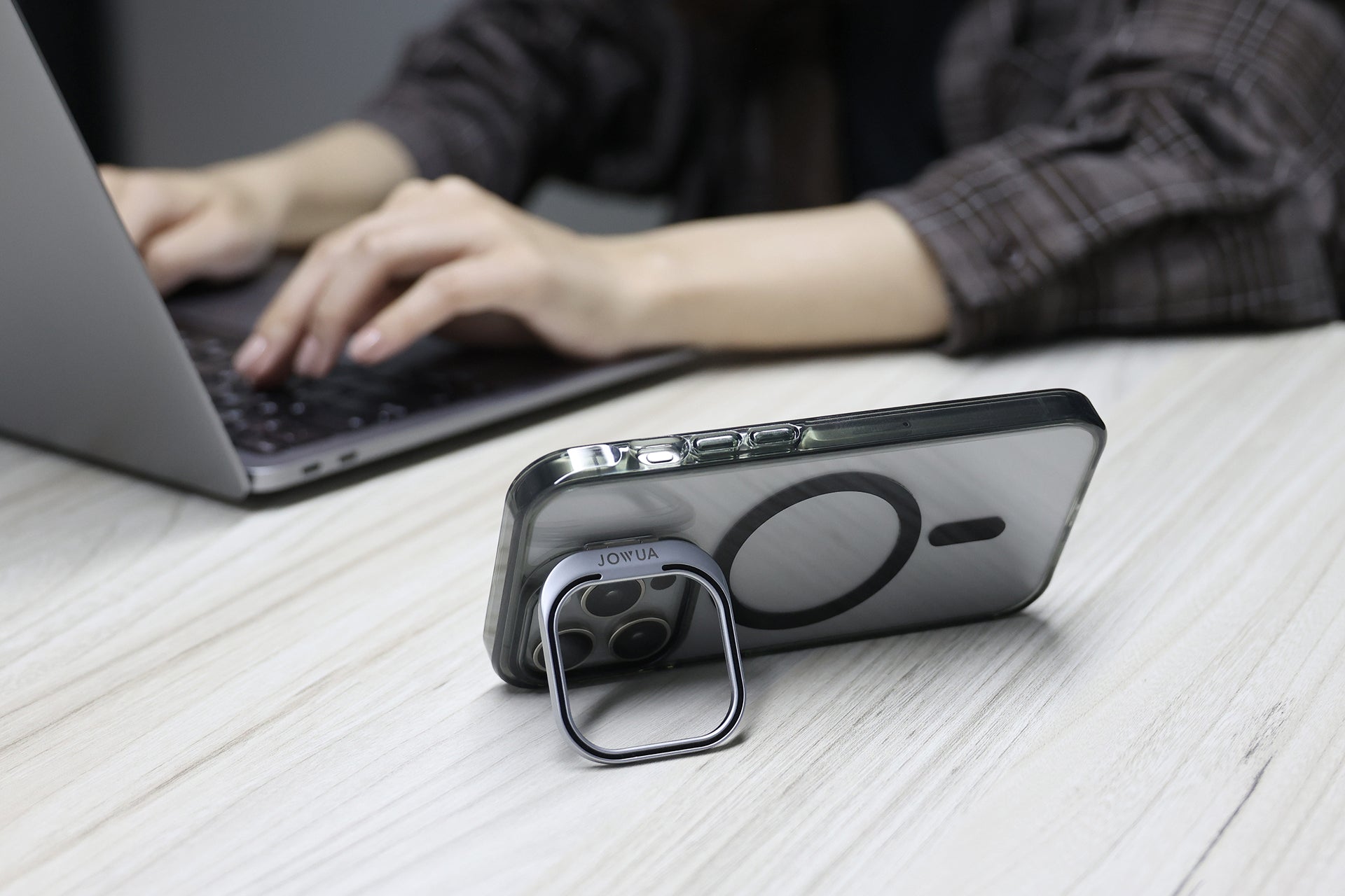 一個人在一張輕木桌上用筆記型電腦打字,而一隻裝有 JOWUA Taiwan MagSafe 透明保護殼 (iPhone 15/14/13 系列) 的 iPhone 就直立在旁邊。只有這個人的手和部分手臂可見。.