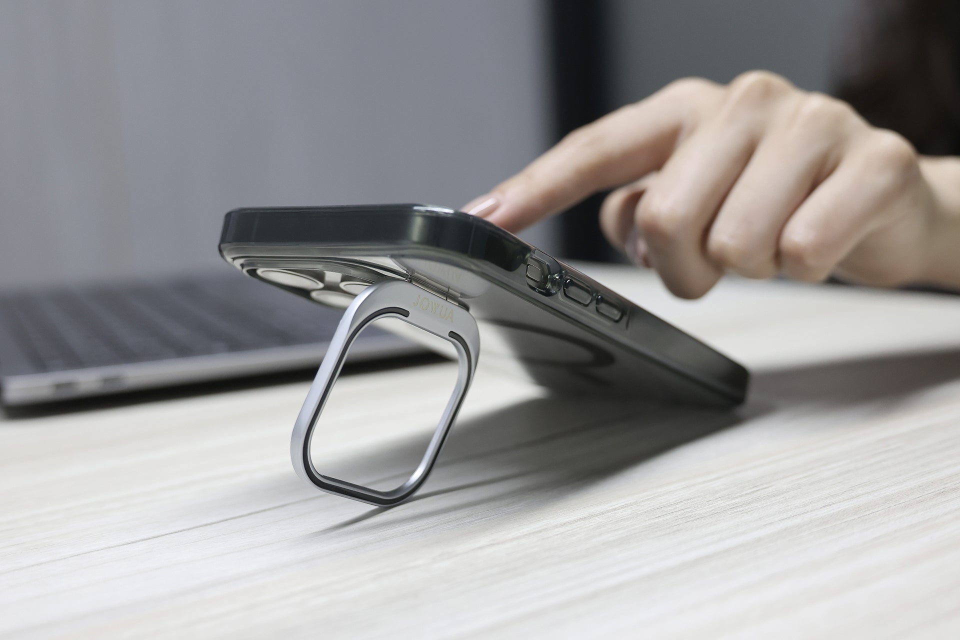 一隻手使用支撐在木質表面上、裝有 JOWUA Taiwan MagSafe 透明支架保護殼 (iPhone 15/14/13 系列) 的智慧型手機,背景則是一台筆記型電腦和其他 MagSafe 磁吸配件。.